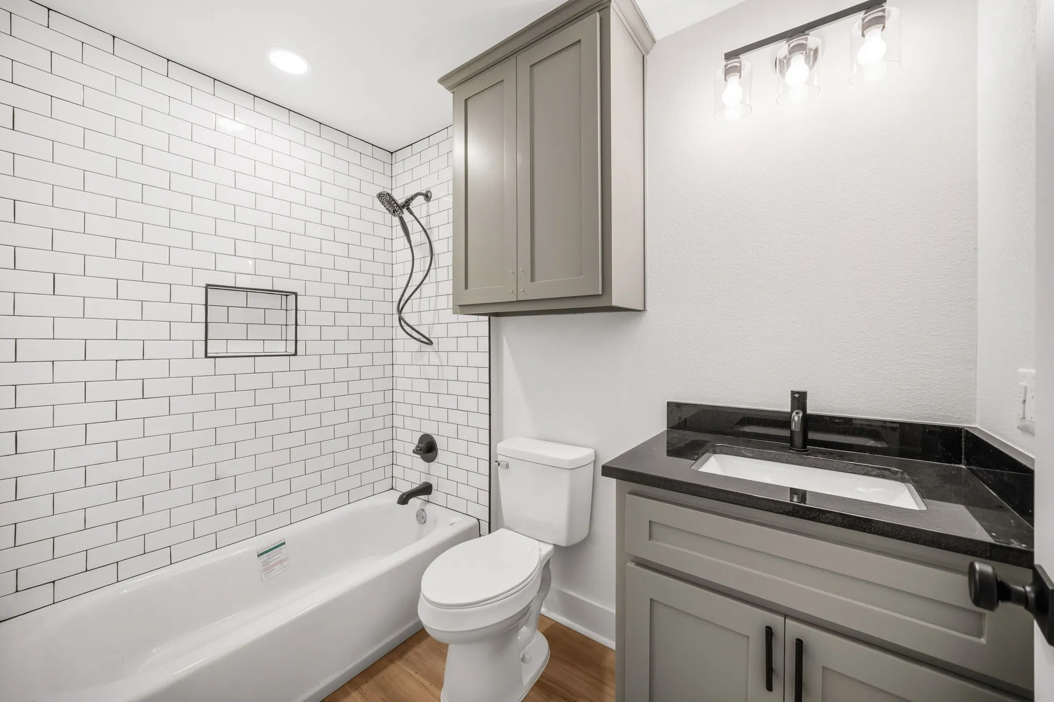 Bathroom featuring a white subway tile shower surround with a built-in niche, matte black fixtures, a gray vanity with a dark countertop, and wood-finish flooring