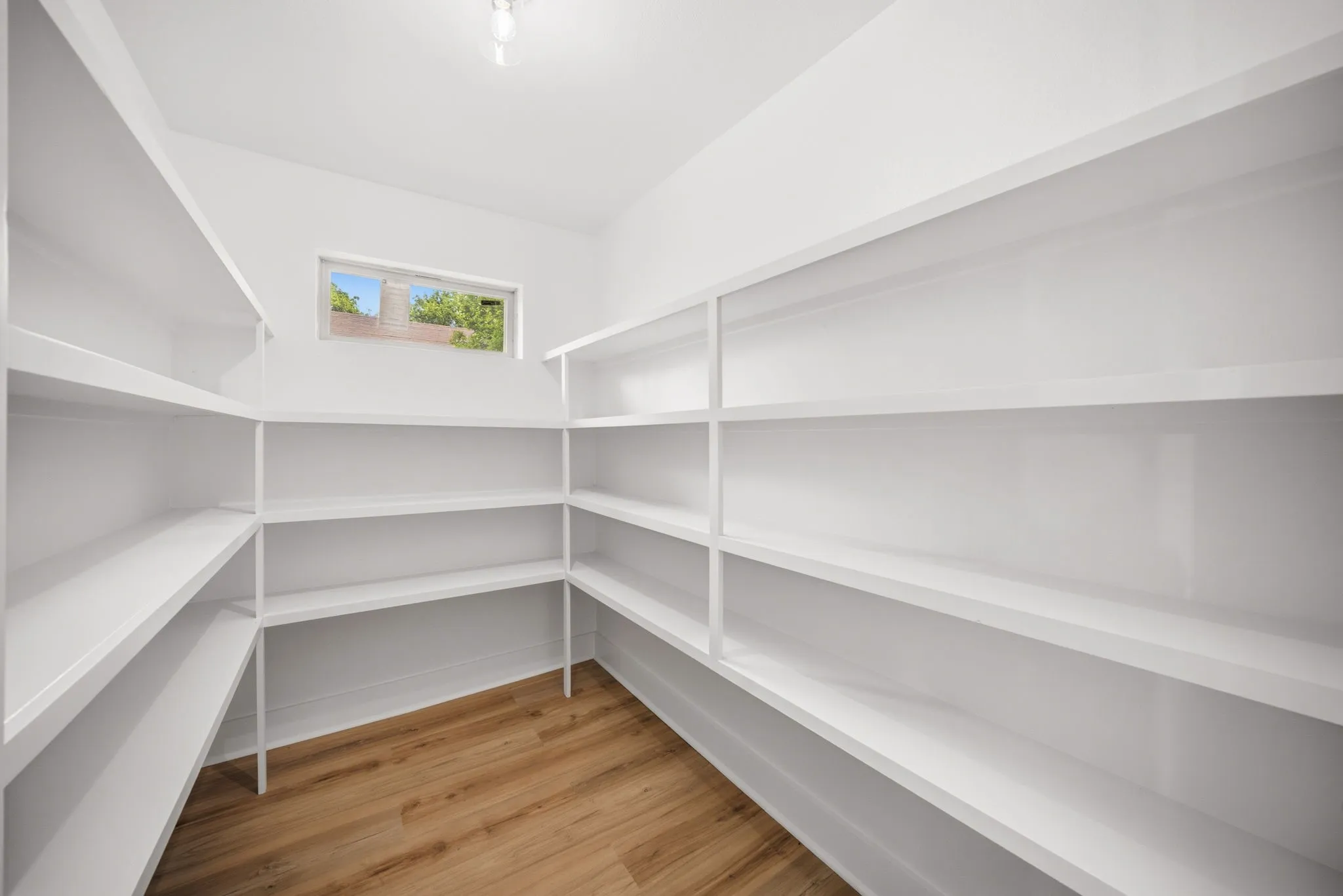 Spacious pantry featuring extensive built-in shelving, wood-finish flooring, a window providing natural illumination, and a ceiling-mounted light fixture