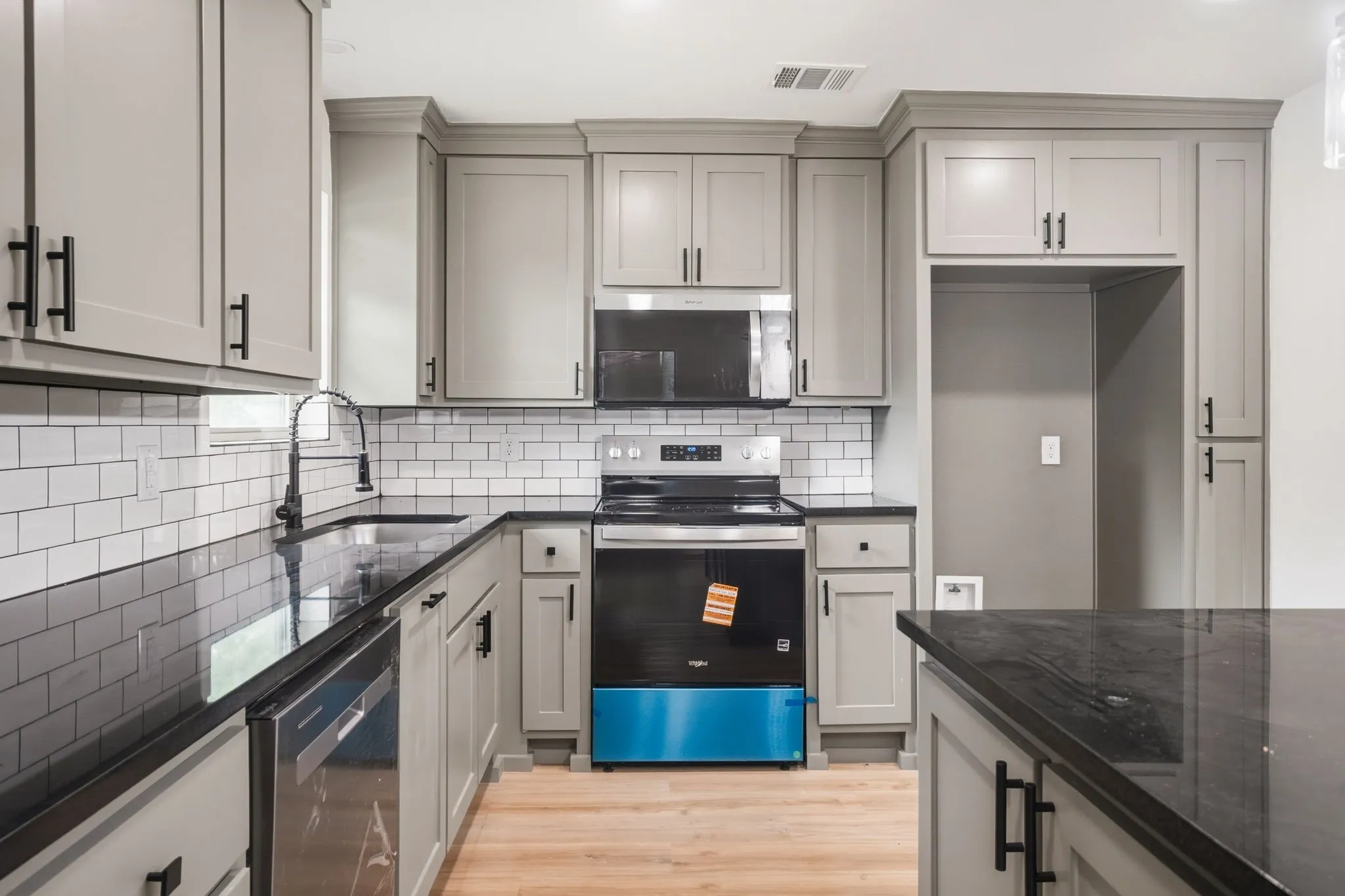 Kitchen featuring light gray cabinetry with black hardware, dark countertops, white subway tile backsplash, stainless steel appliances, and wood-finish flooring