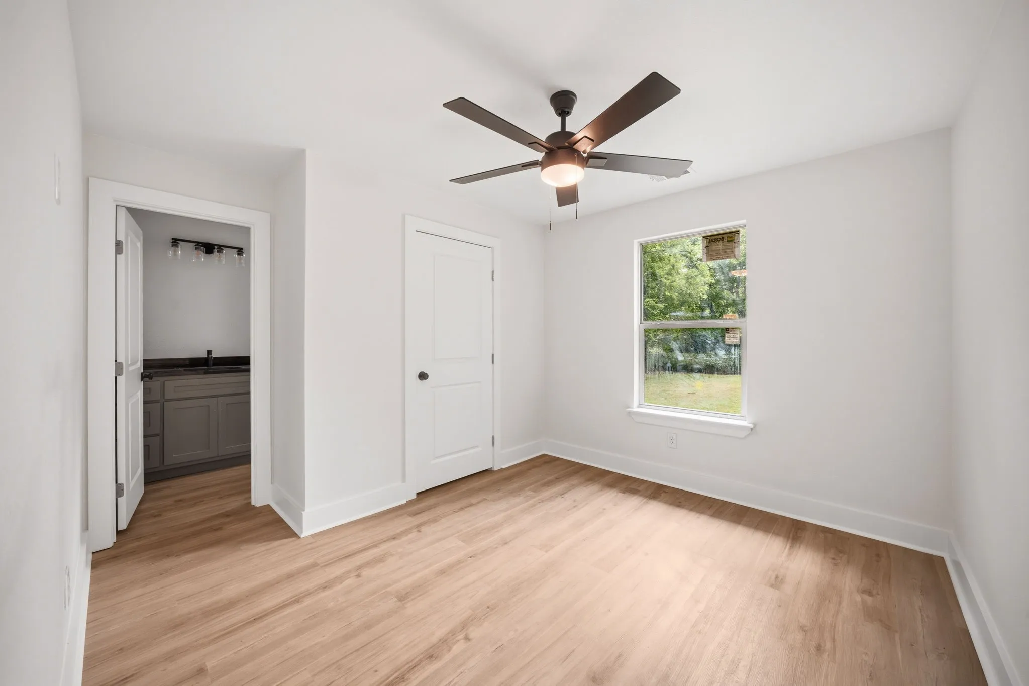 Room featuring wood-finish flooring, a ceiling fan with integrated lighting, and a window overlooking exterior greenery