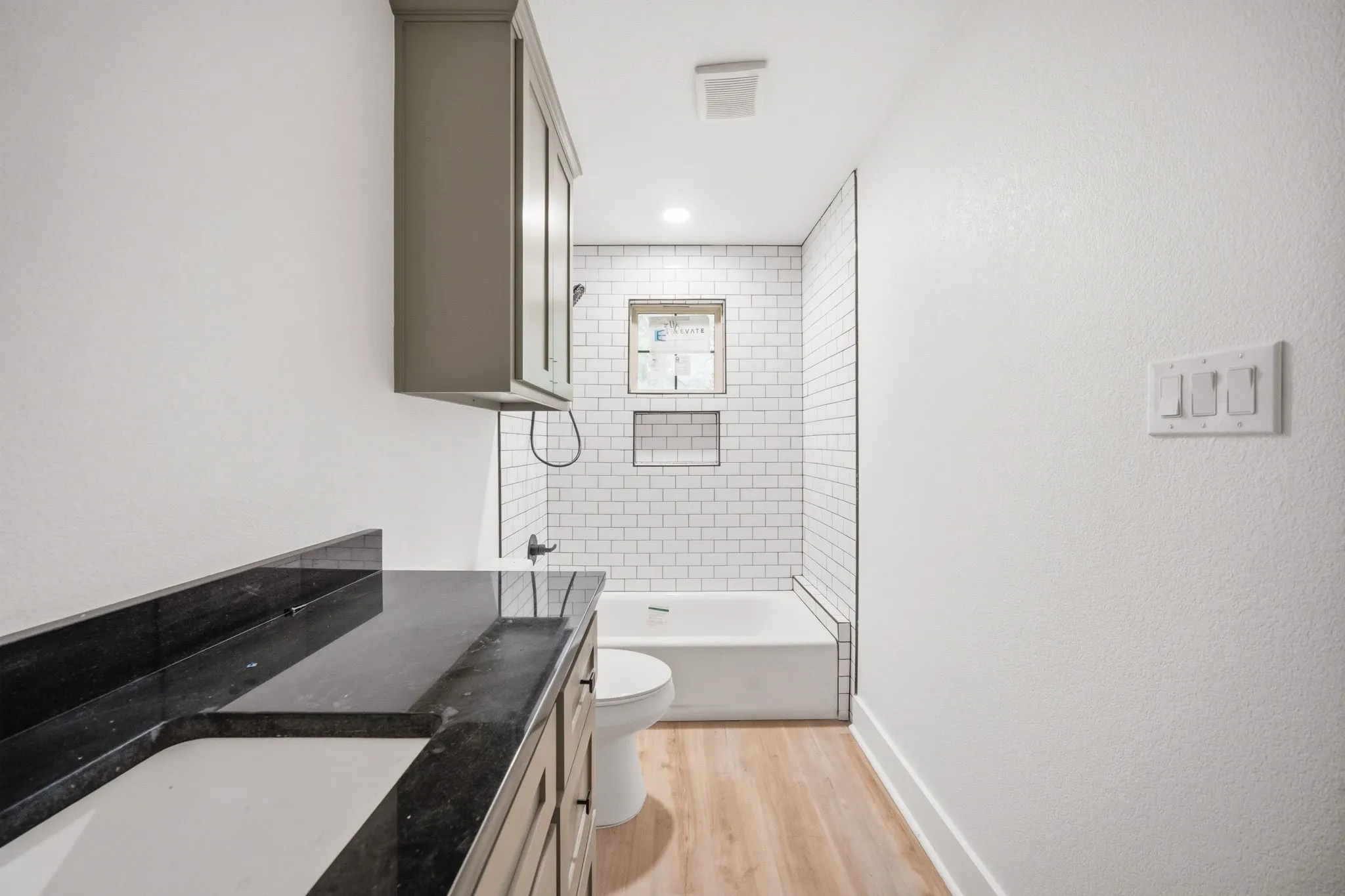 Bathroom featuring a dark countertop vanity with undermount sink, overhead cabinetry, subway tile shower surround with built-in niches, and wood-finish flooring