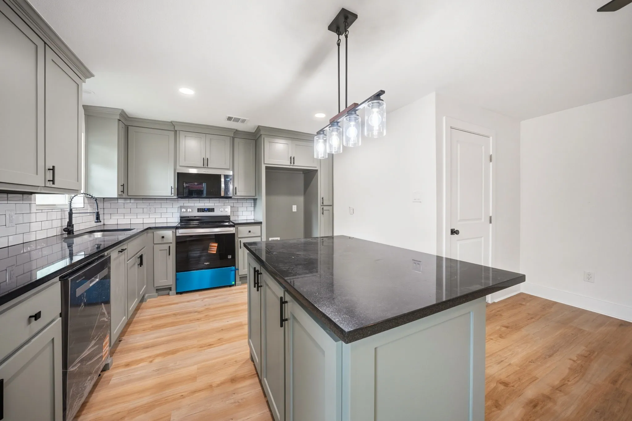Modern kitchen featuring light wood-finish flooring, light grey cabinetry, dark countertops, white subway tile backsplash, and a central island with an overhead linear pendant light fixture