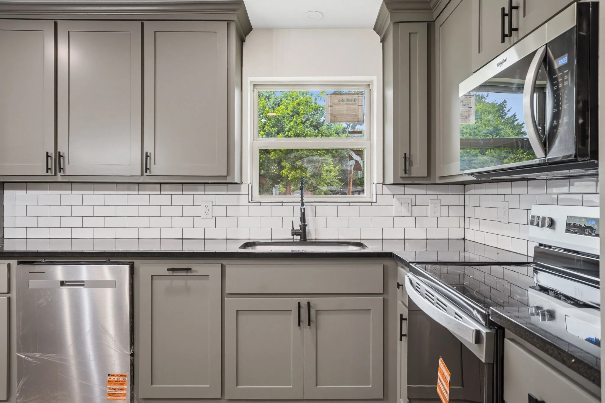 Modern kitchen featuring gray shaker cabinetry, black countertops, a white subway tile backsplash, stainless steel appliances, and a window above the sink