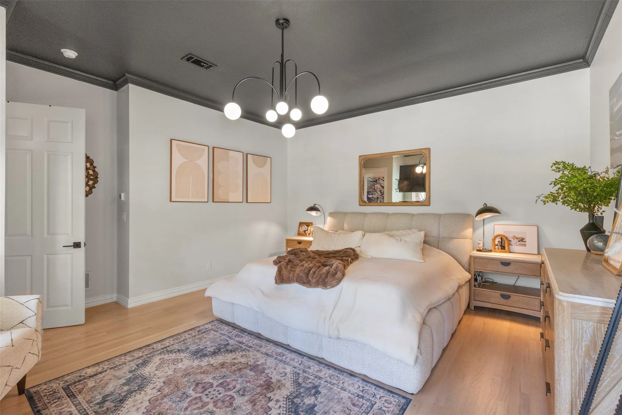 Designer bedroom retreat featuring white oak flooring, a dramatic dark ceiling with matching crown molding, modern statement lighting, and warm natural wood furnishings—creating a sophisticated, elevated space with a boutique-luxury feel.