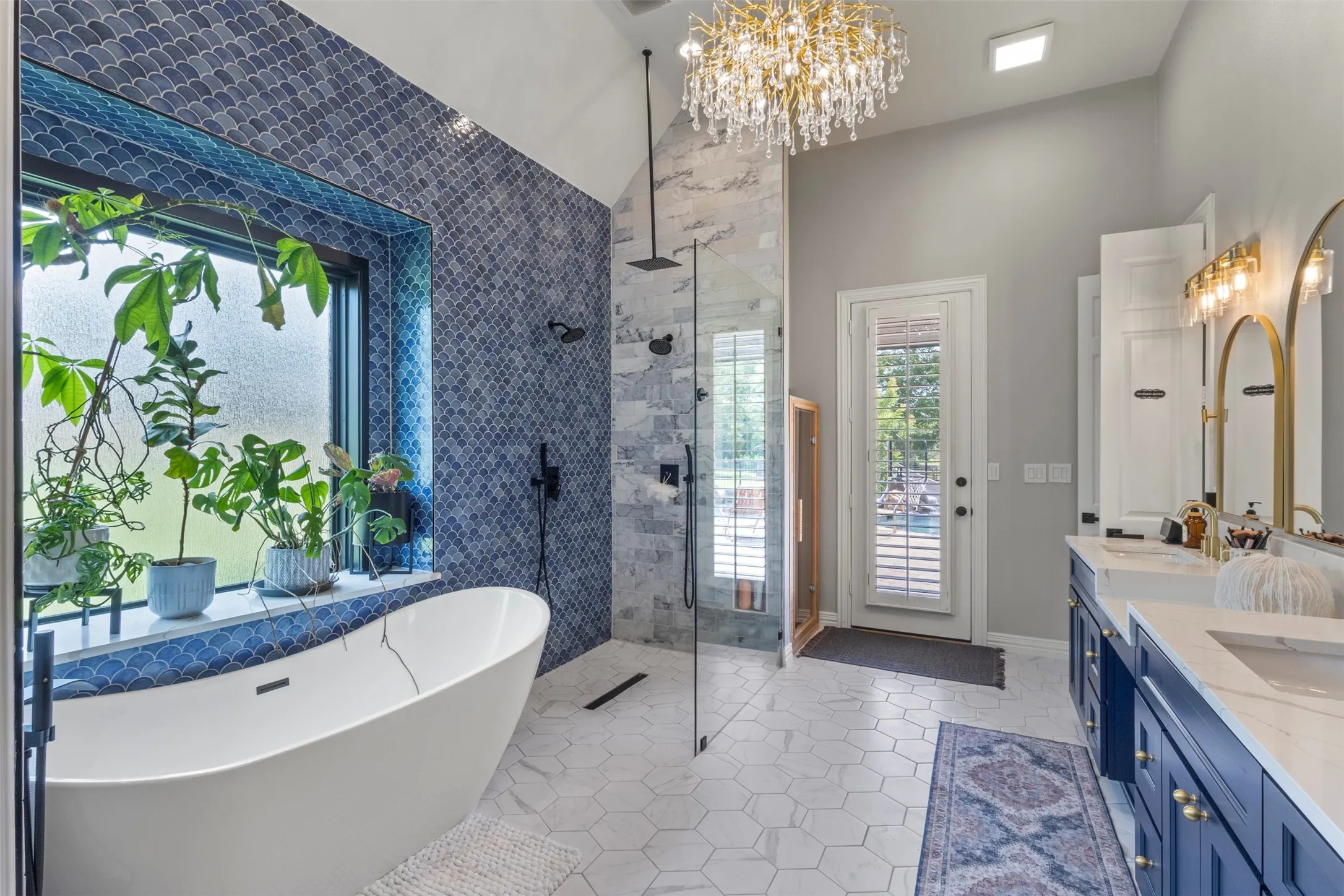 Spa-inspired primary bathroom fully renovated with 12’ ceilings, floor-to-ceiling tile, a custom frameless glass shower, standalone soaking tub, Calacatta quartz countertops, and 16’ of Sherwin-Williams Naval custom cabinetry with gold hardware and a statement teardrop glass chandelier.