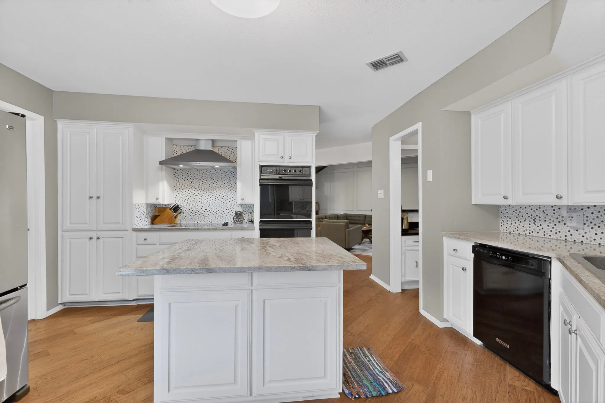 Open-concept kitchen featuring an island with a light-toned countertop, white cabinetry, wood-finish flooring, stainless steel range hood, and integrated black appliances