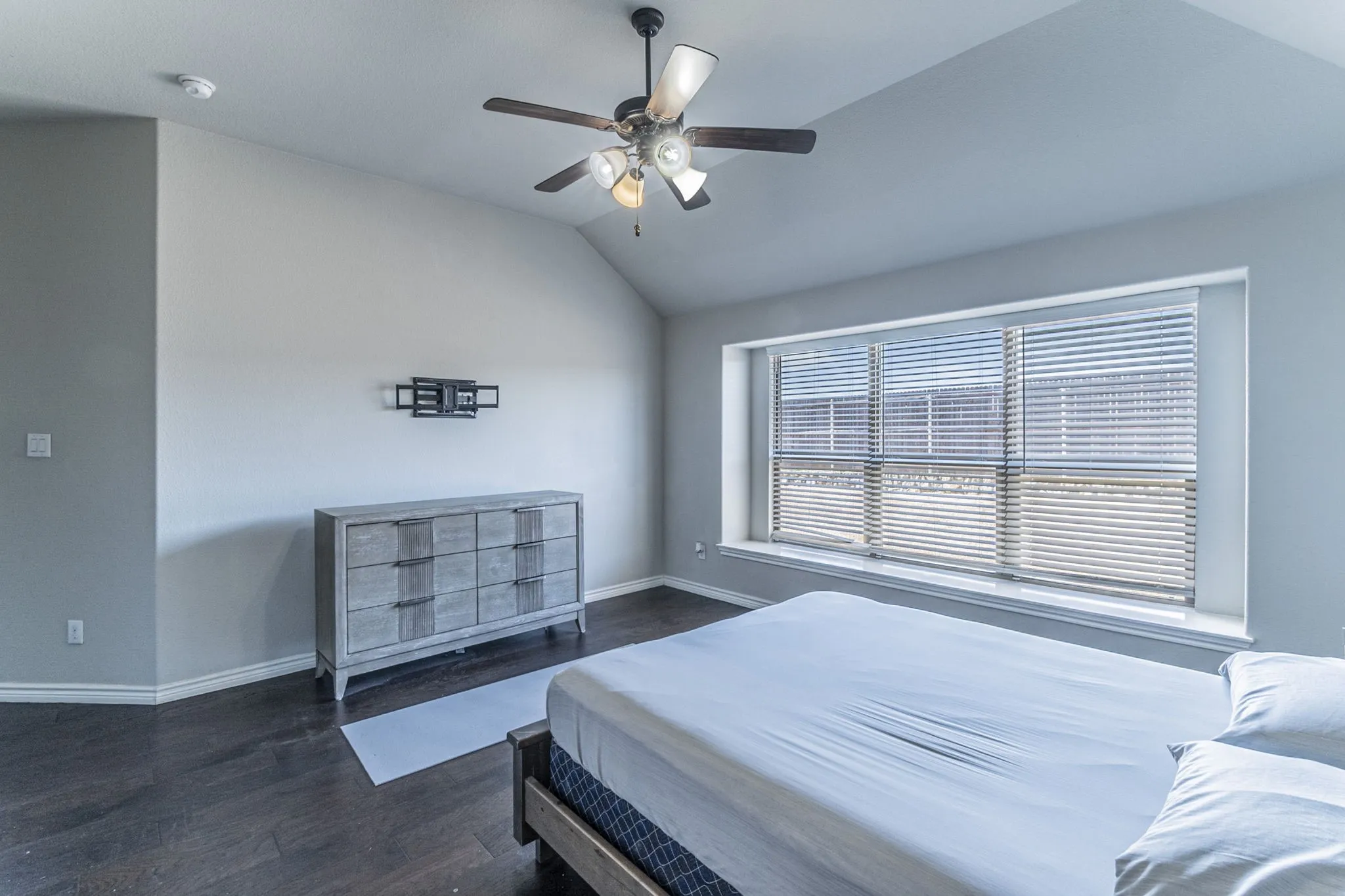 Spacious room with dark wood-finish flooring, vaulted ceiling, and a large window featuring blinds
