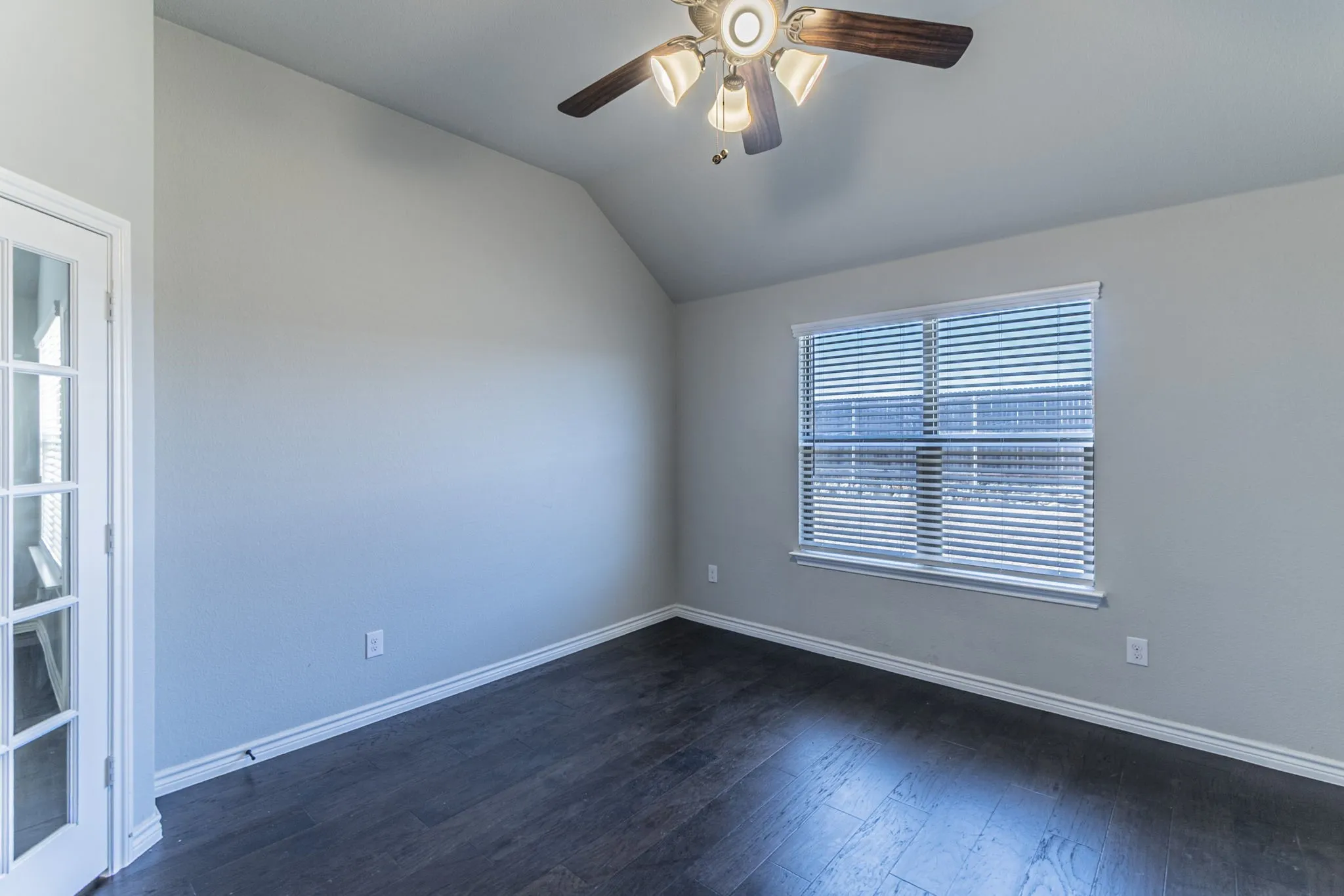Vaulted ceiling accentuates this room featuring dark wood-finish flooring, a window with blinds, and white trim