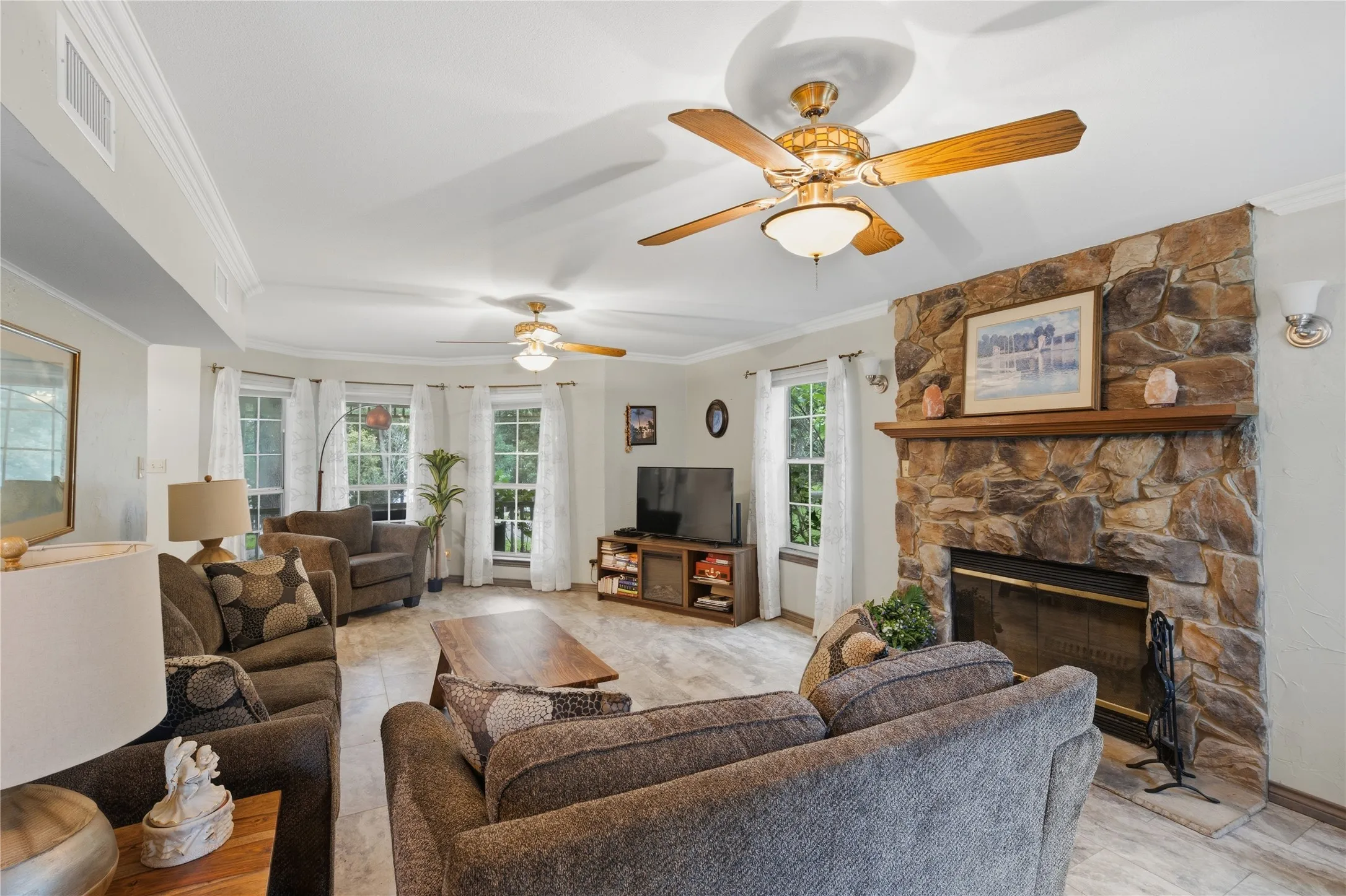Spacious living area featuring a stone-clad fireplace with a wood mantel, multiple windows, tile flooring, and crown molding