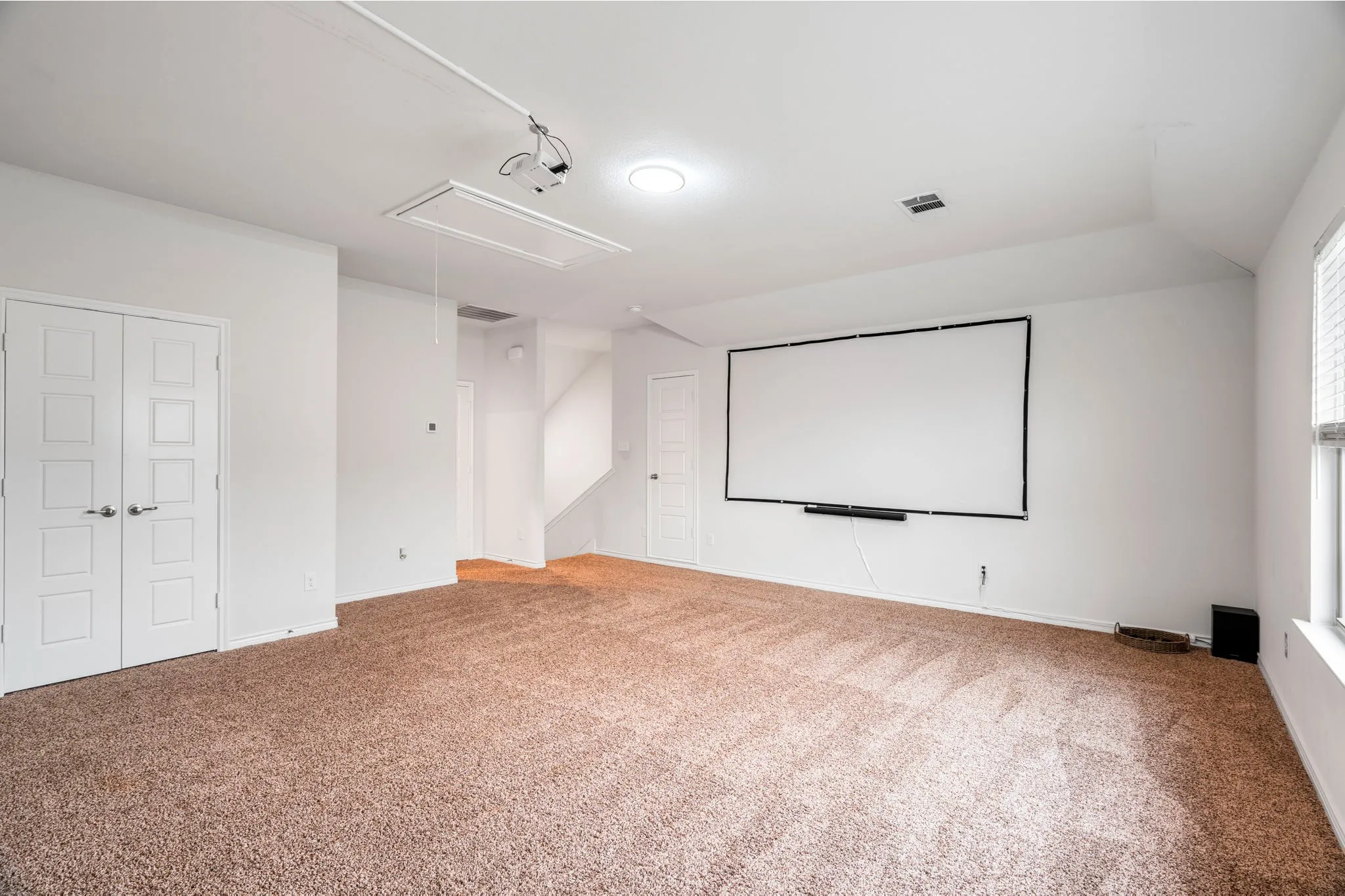 Spacious room featuring a mounted projector and screen, recessed lighting, neutral tone carpeting, and white paneled double doors
