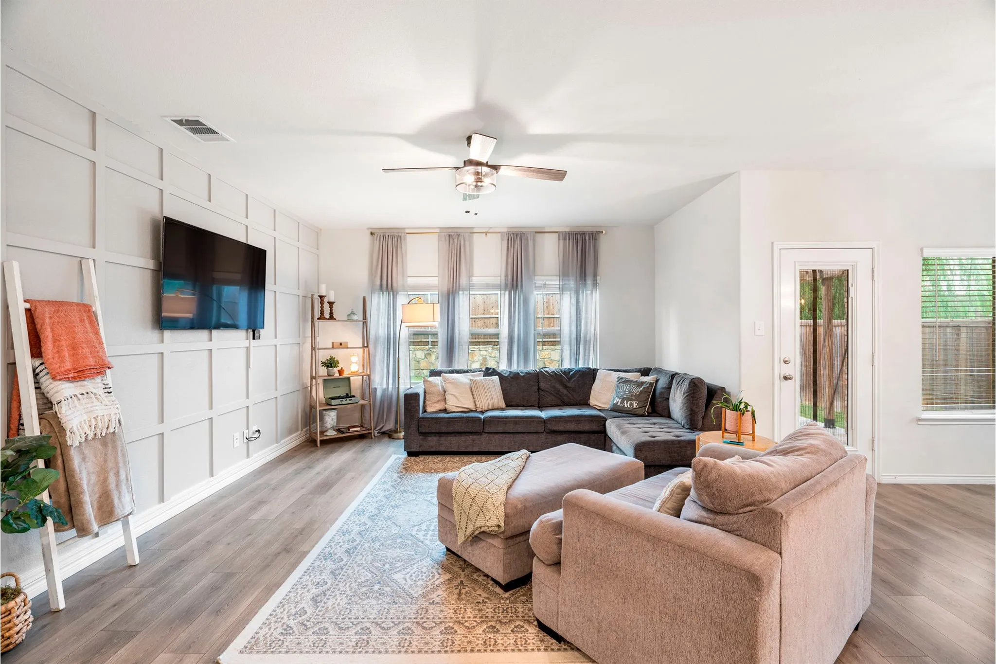 Spacious living area featuring wood-finish flooring, a decorative accent wall with paneling, a ceiling fan, and multiple windows for natural light