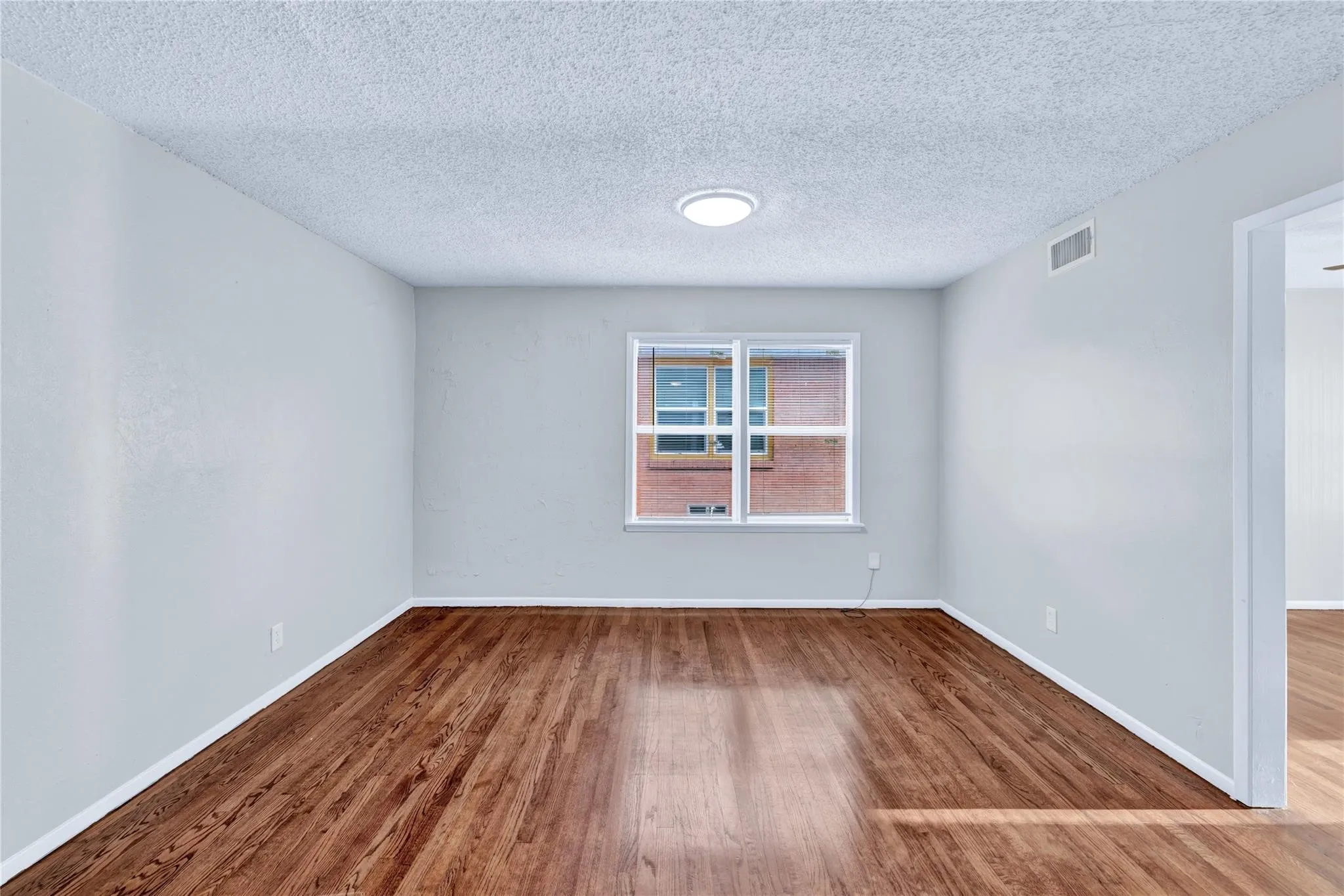 Spacious room featuring wood-finish flooring, light gray walls, a large window, and a ceiling-mounted light fixture