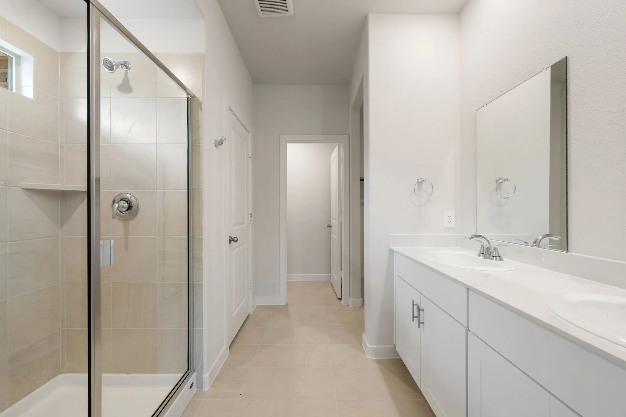 Bathroom featuring a dual-sink vanity with white cabinetry, a walk-in shower with tile surround and glass enclosure, and light-toned tile flooring