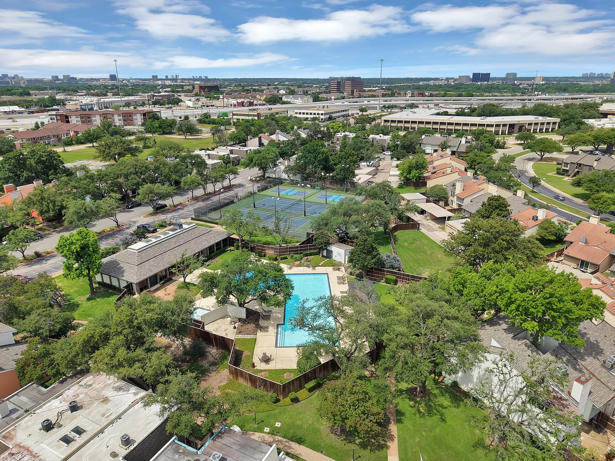 Community amenities featuring a central swimming pool with surrounding lounge areas, two tennis courts, and a clubhouse building, all set amidst mature trees and residential structures