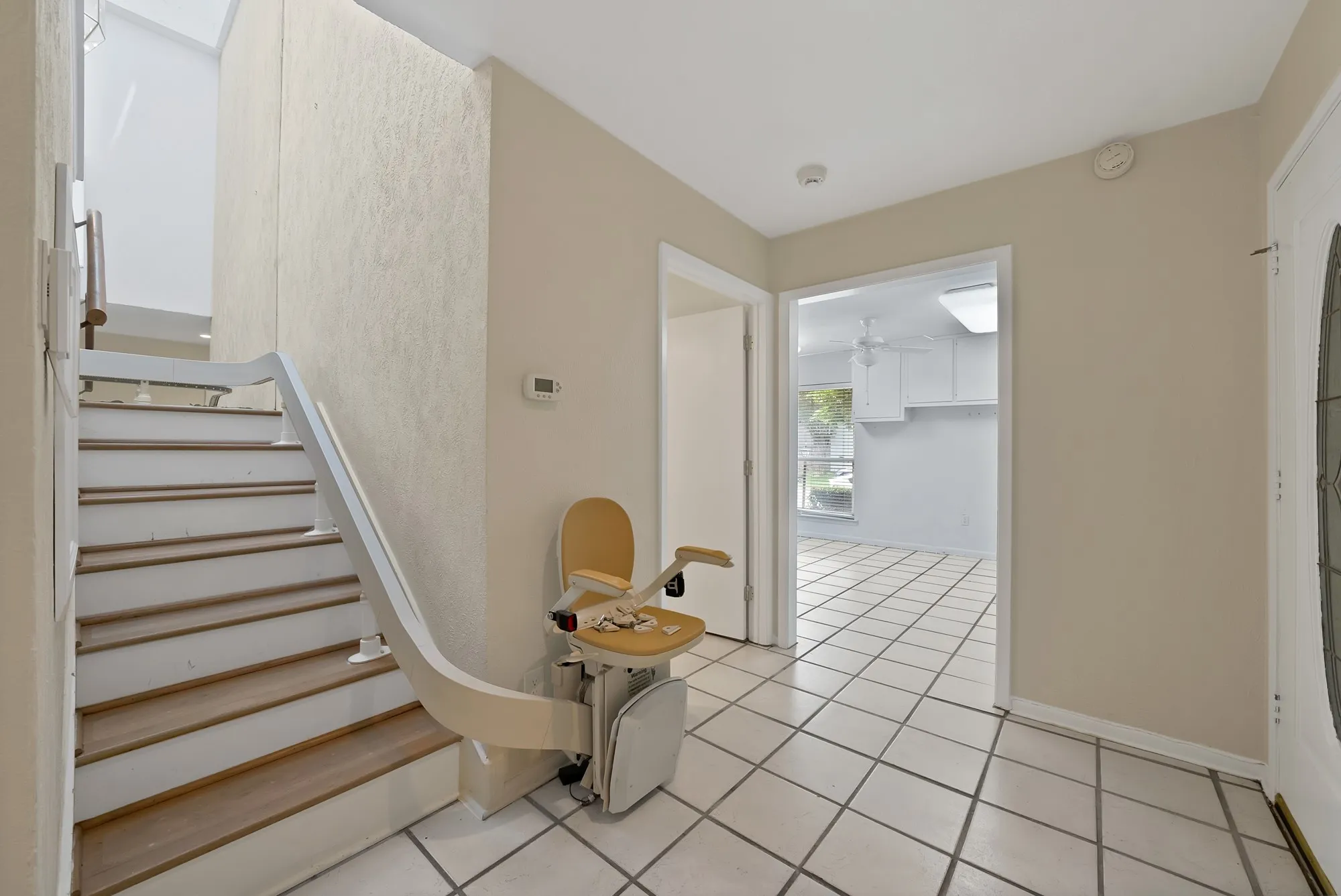 Entry foyer featuring white square tile flooring with contrasting grout, a wood-finish staircase with white risers and railing, an integrated stair lift system, and a front entry door with an oval glass insert