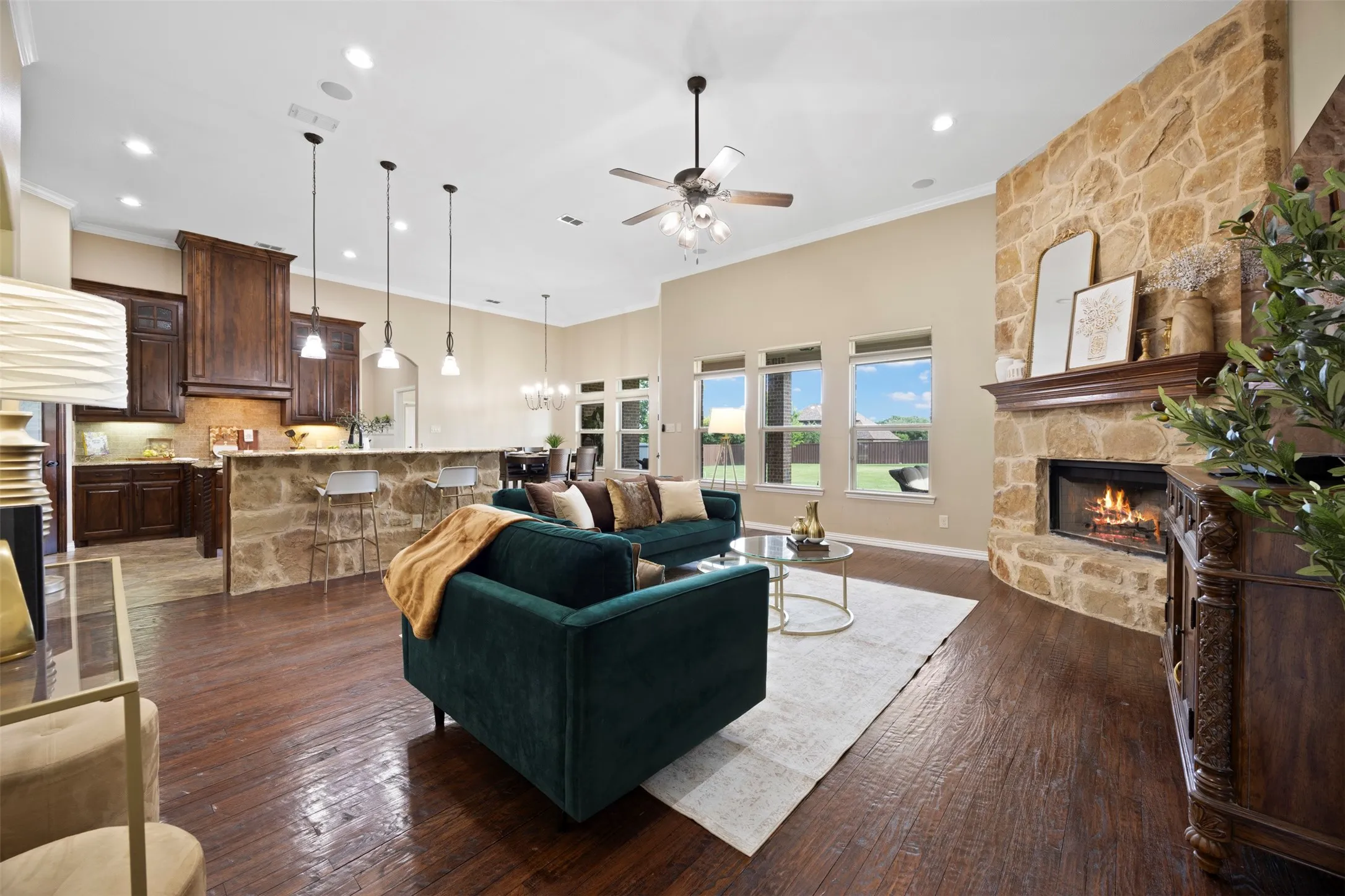 Open-concept living space featuring wood-finish flooring, a stone-clad fireplace with a wood mantel, and multiple windows offering backyard views