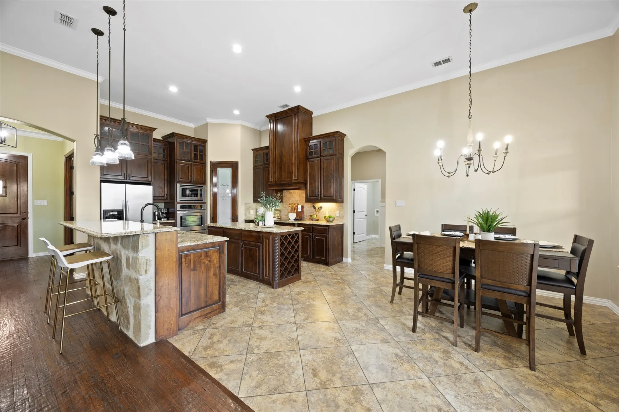 Spacious kitchen and dining area featuring rich wood cabinetry, stainless steel appliances, stone-clad island with seating, pendant lighting, and ceramic tile flooring