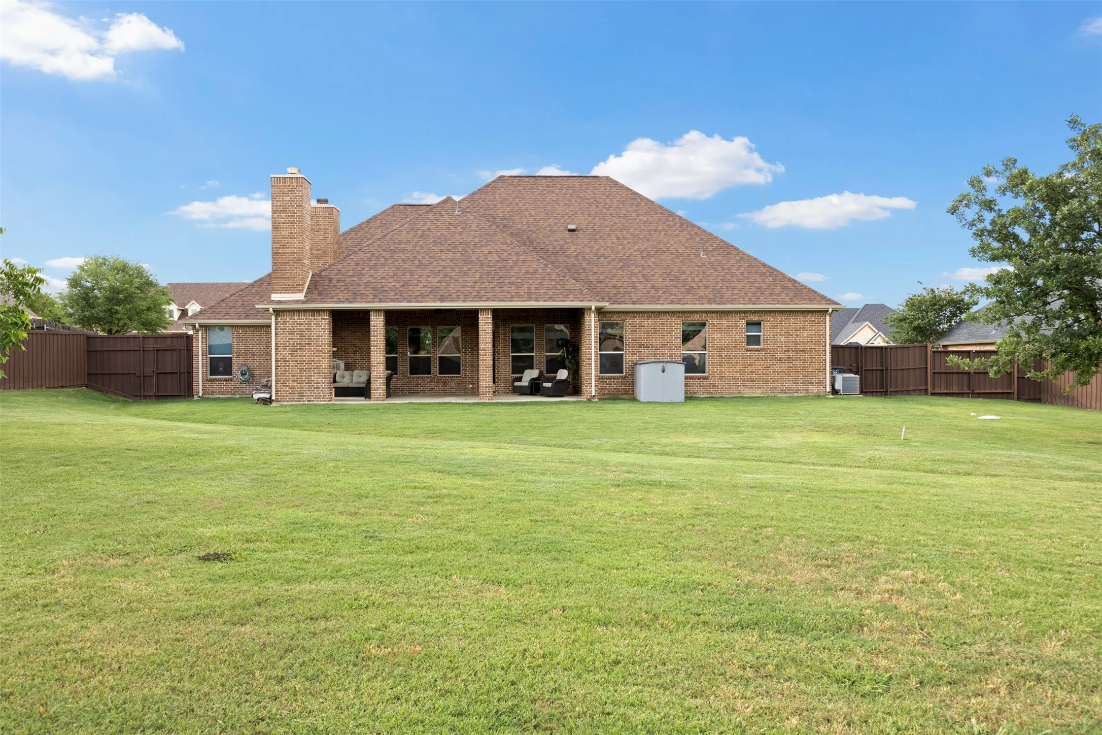 Expansive fenced backyard featuring a substantial covered patio, a brick exterior with multiple windows, and a brown shingle roof