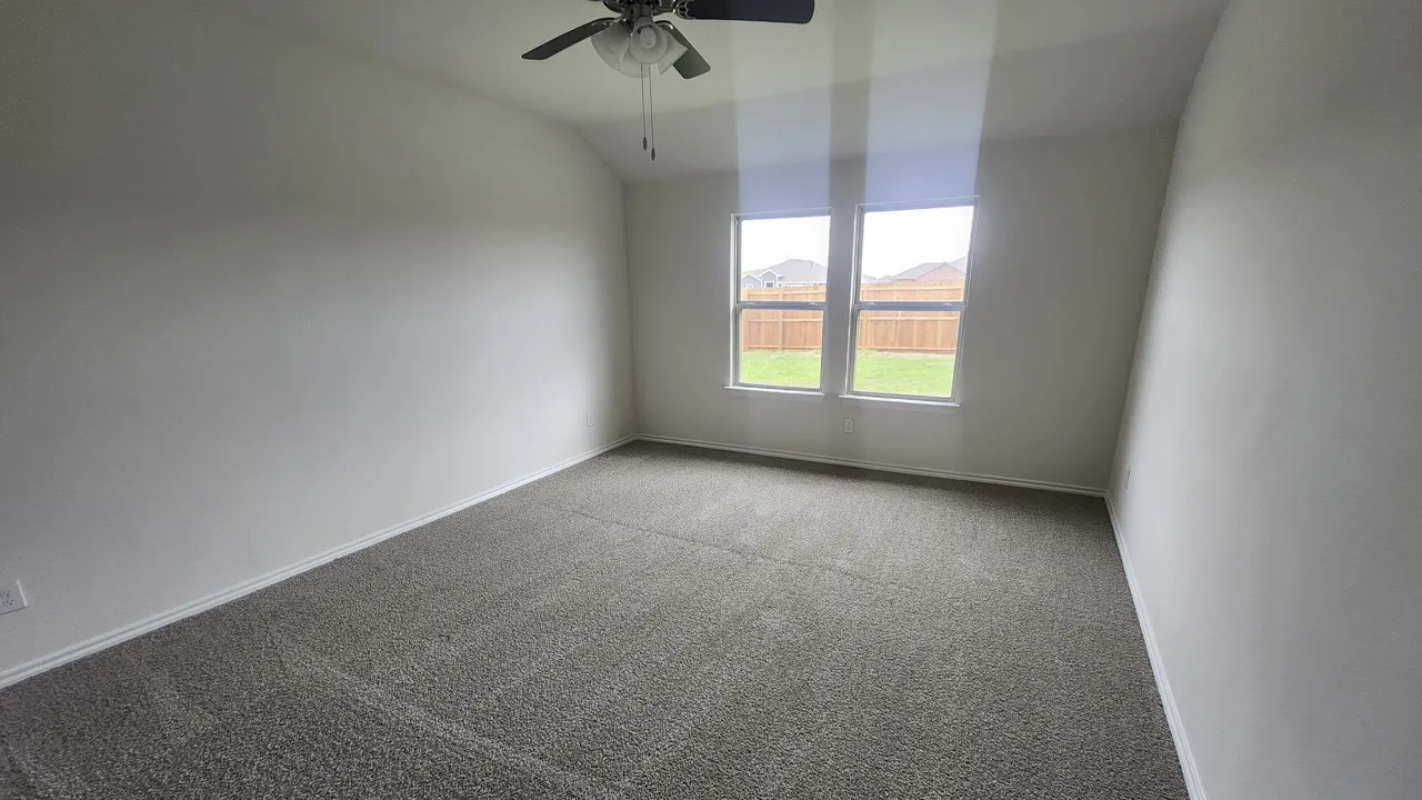 Spacious room featuring neutral carpeting, a ceiling fan, and two large windows offering views of the fenced backyard
