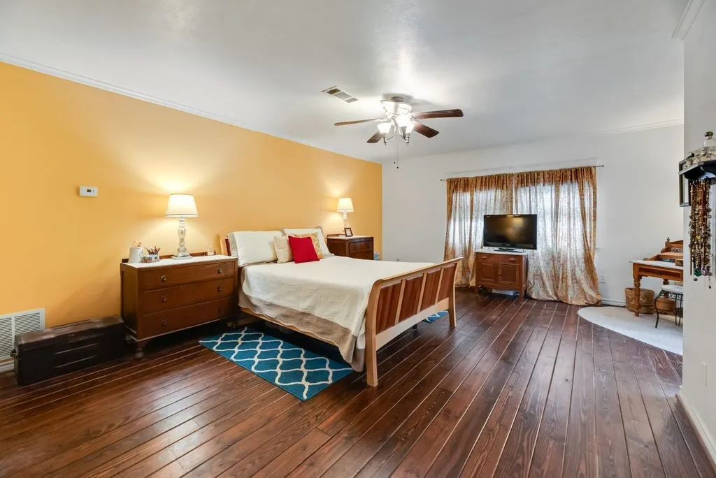 Spacious room featuring rich wood-finish flooring, a ceiling fan with integrated lighting, and a window with curtain rods