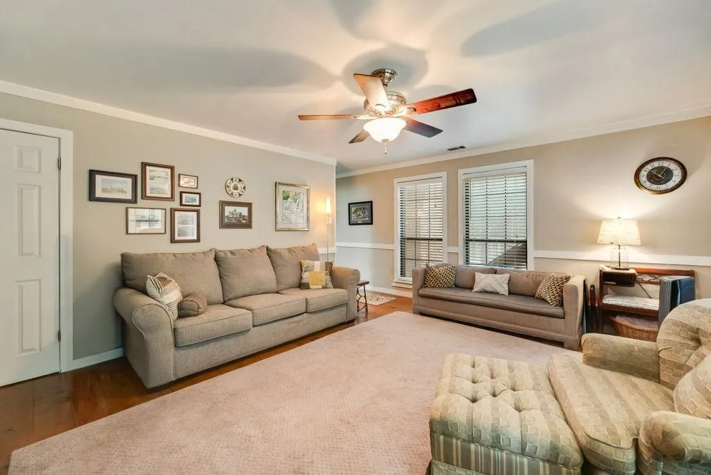 Spacious living area featuring warm wood-finish flooring, a neutral color palette, twin windows with blinds, white crown molding, and a ceiling fan with integrated lighting