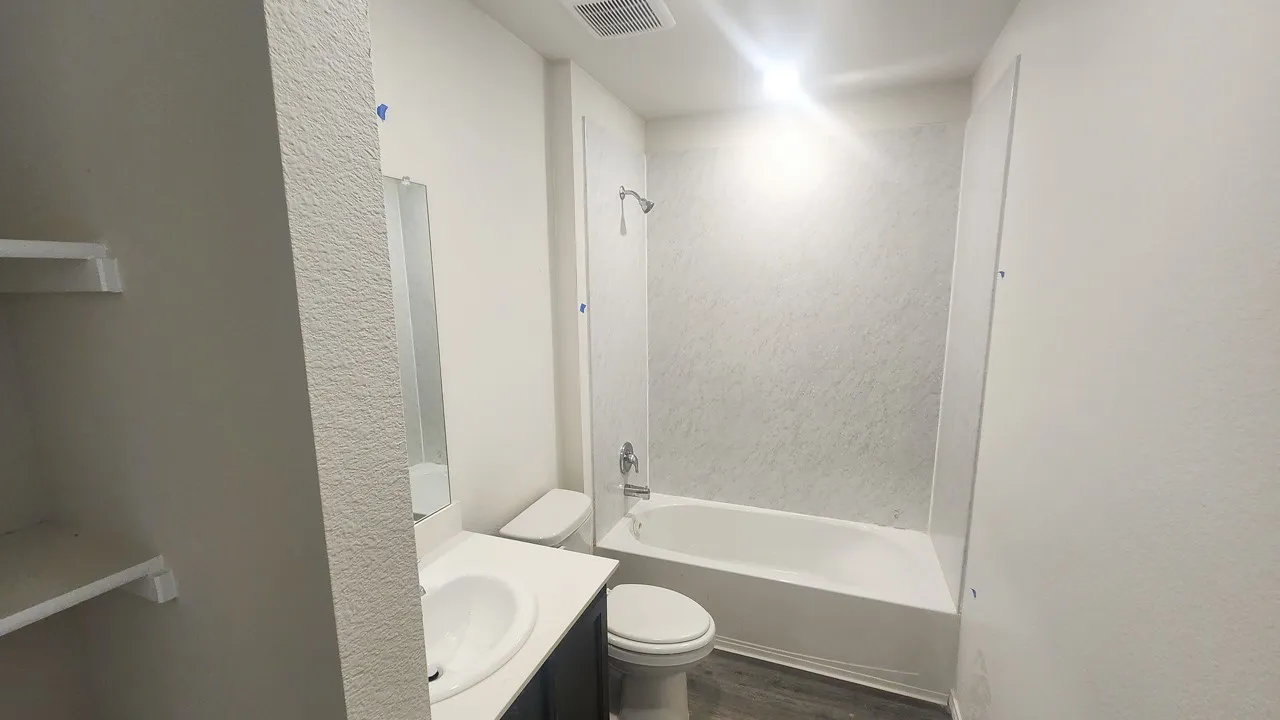 Full bathroom featuring a white porcelain tub with integrated shower, vanity with an undermount sink and dark cabinetry, a wall-mounted mirror, and wood-finish flooring