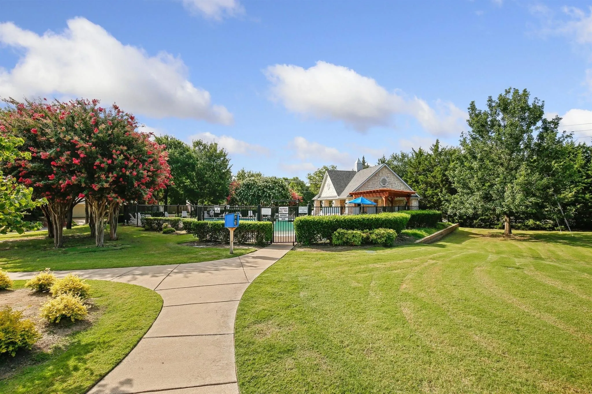 Manicured grounds featuring a concrete pathway, a swimming pool enclosure, a clubhouse with an attached pergola, mature trees, and flowering shrubs
