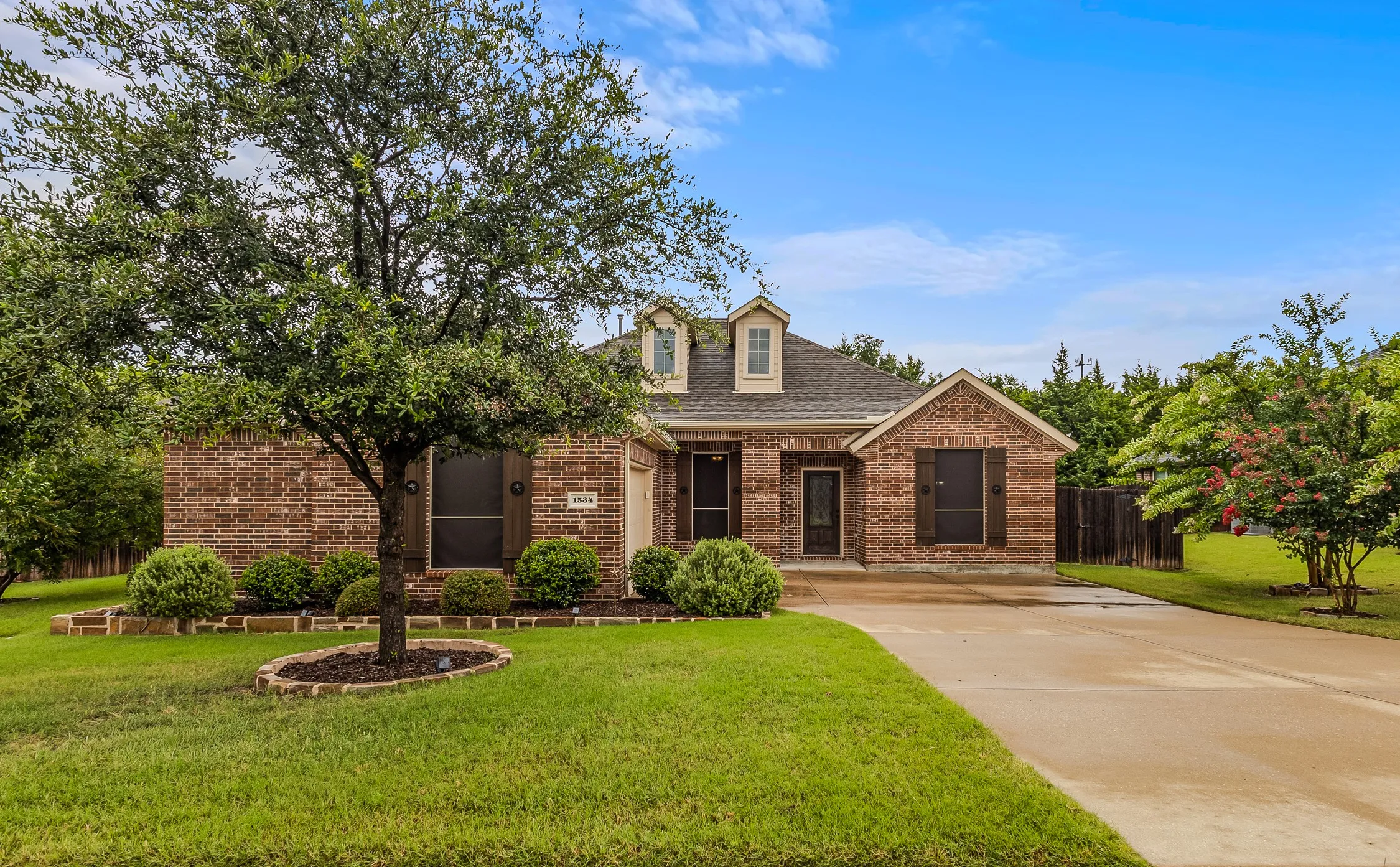 Brick exterior featuring dormer windows, a paved driveway, mature front yard tree, and established shrubbery