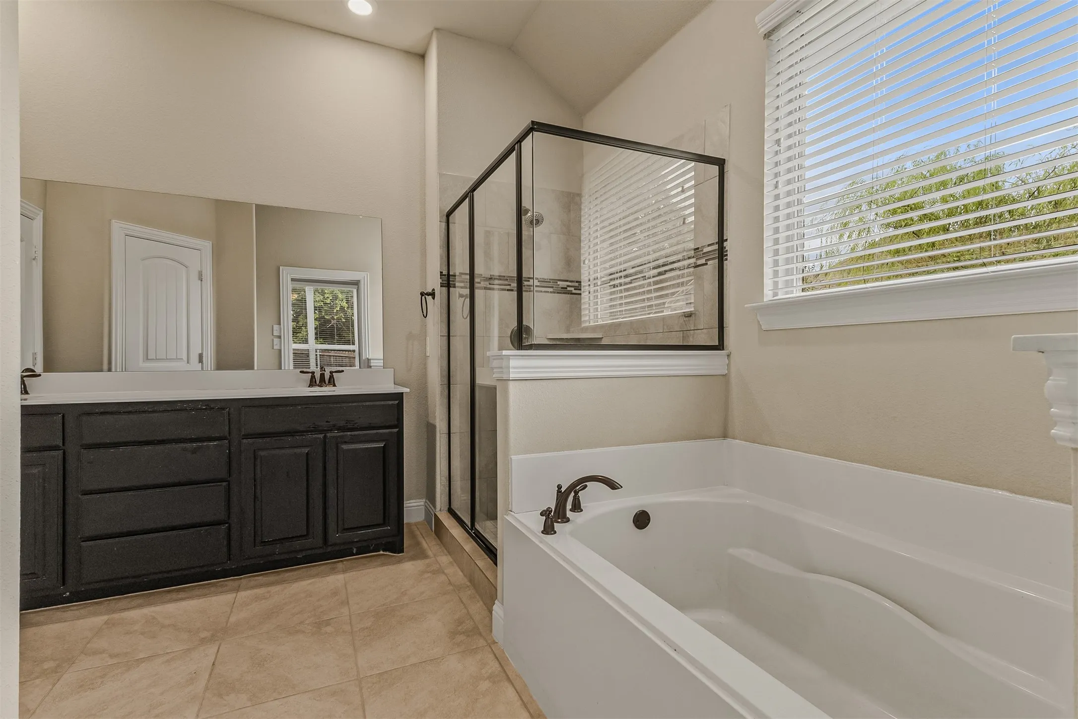Bathroom featuring a dual vanity with dark cabinetry, integrated soaking tub, separate glass-enclosed shower, and tiled flooring