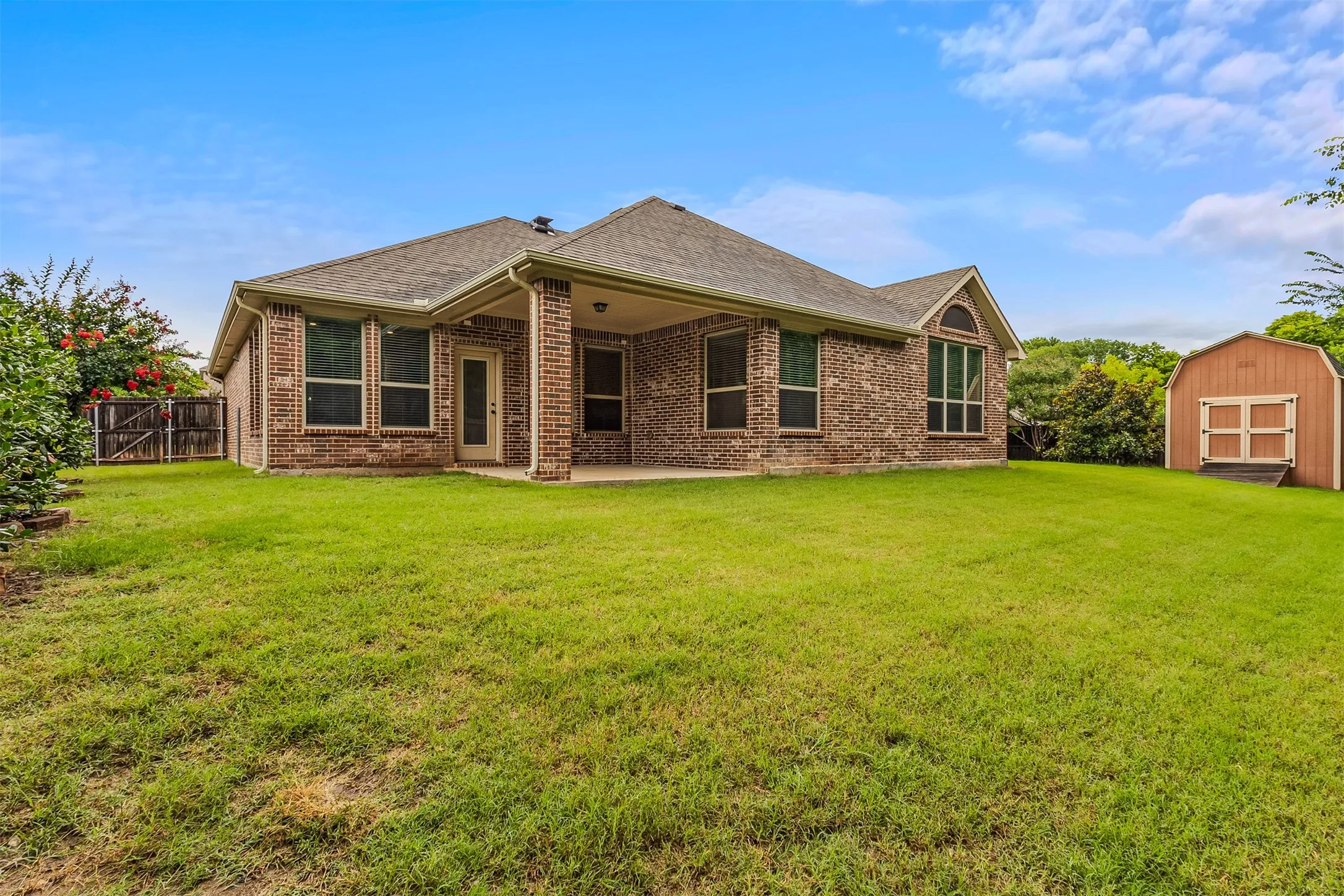 GREENBELT VIEWS. Expansive backyard featuring a brick exterior, covered patio, multiple windows, a gambrel-roofed storage shed, and perimeter fencing