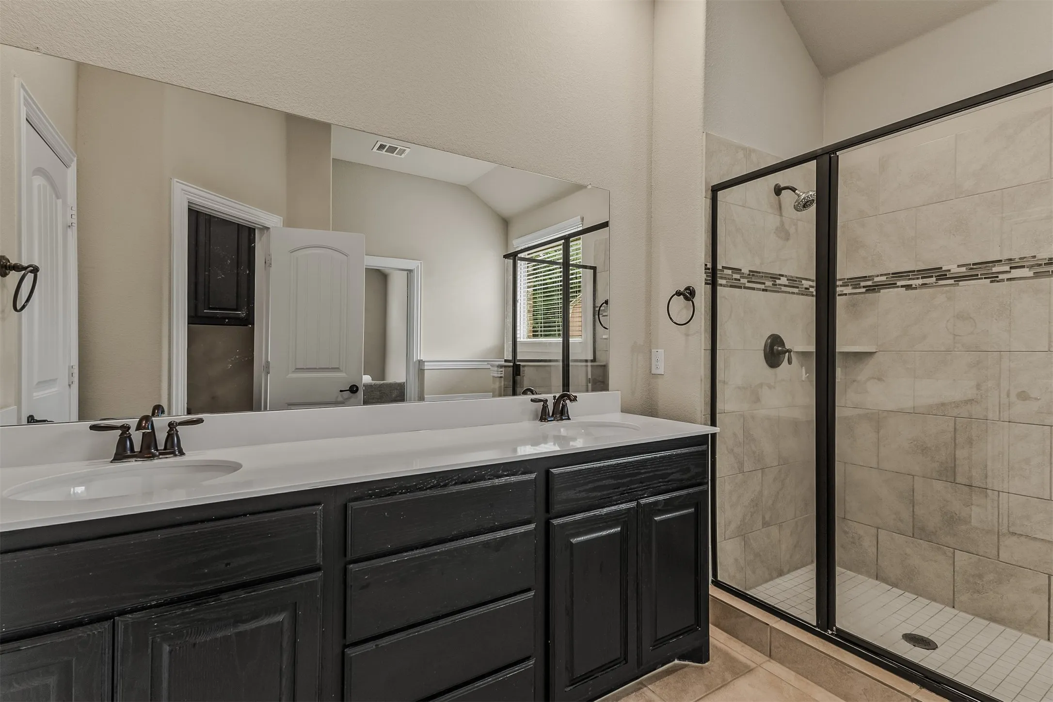 Dual vanity with undermount sinks and dark cabinetry, quartz countertop, large wall-mounted mirror, glass-enclosed shower with tiled walls and decorative accent band, and bronze fixtures