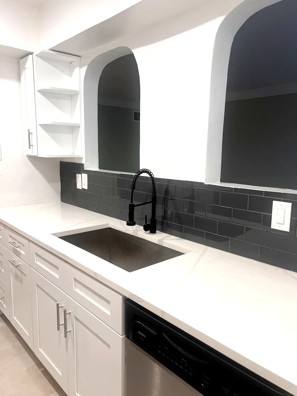 Modern kitchen sink area featuring white shaker cabinetry, light-toned countertops, a dark subway tile backsplash, a matte black faucet, and a stainless steel under-mount sink