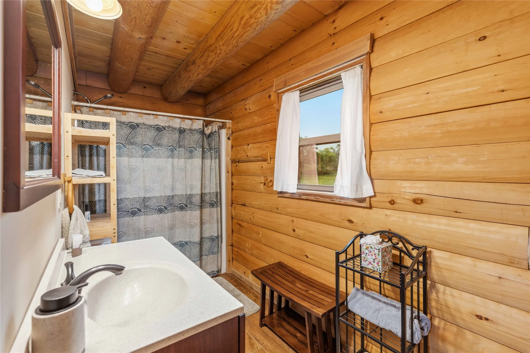Bathroom featuring exposed log walls, a single vanity with a white countertop and dark base, a built-in wooden shelving unit, a window with white curtains, and a shower area