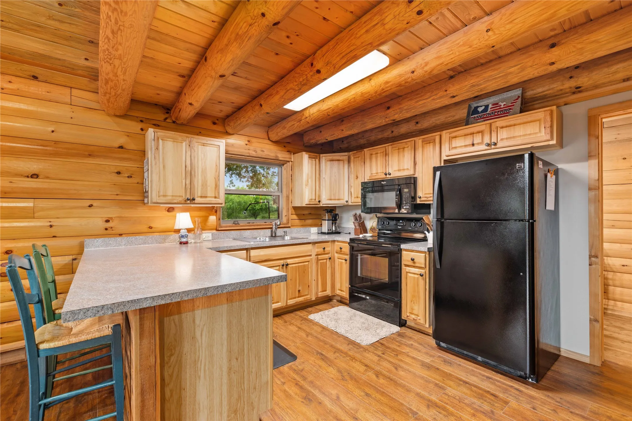 Kitchen featuring log walls and ceiling beams, light wood cabinetry, a built-in breakfast bar with a solid surface countertop, black appliances, and wood-finish flooring