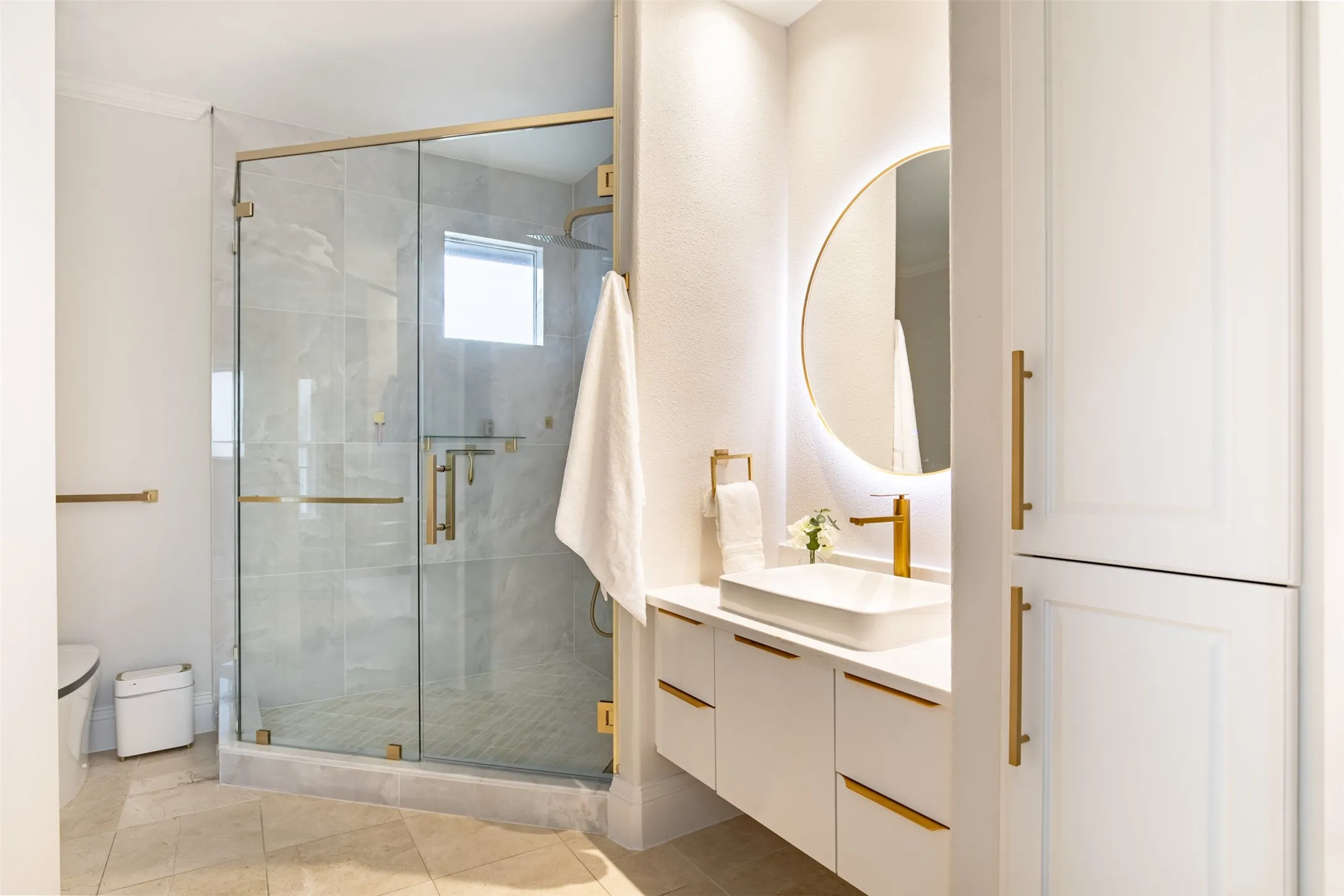 Contemporary bathroom featuring a frameless glass shower enclosure with brass hardware, a floating vanity with an integrated rectangular sink, a circular back-lit mirror, and custom built-in cabinetry with brass pulls