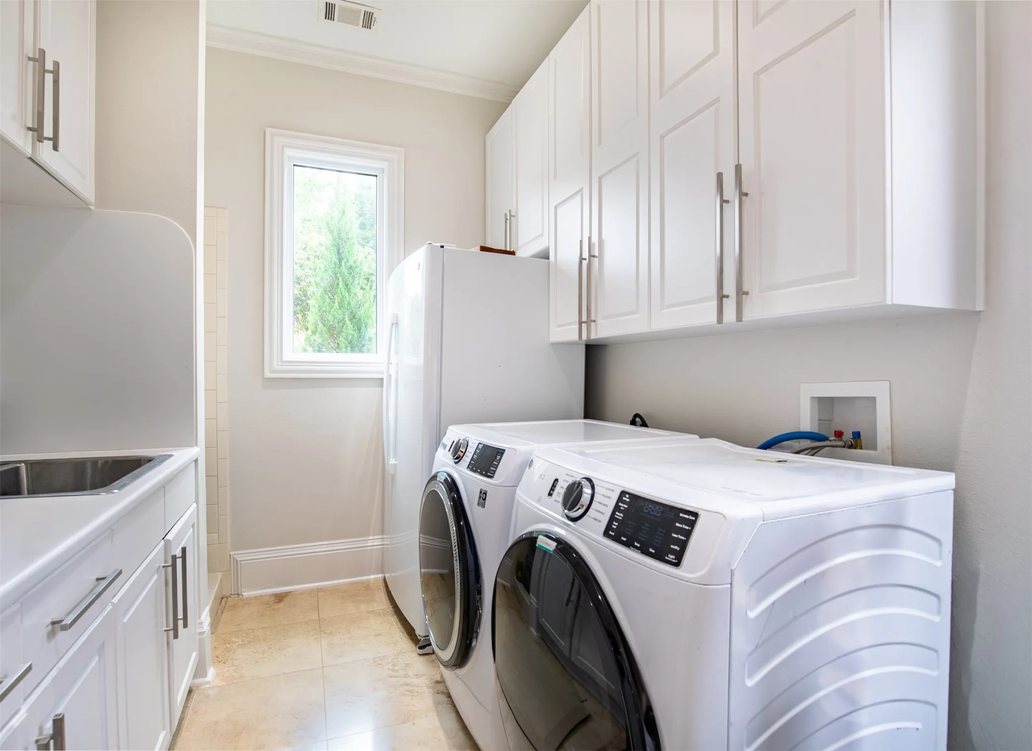 Dedicated utility space featuring white cabinetry with brushed nickel hardware, a stainless steel utility sink, tile flooring, and a single window