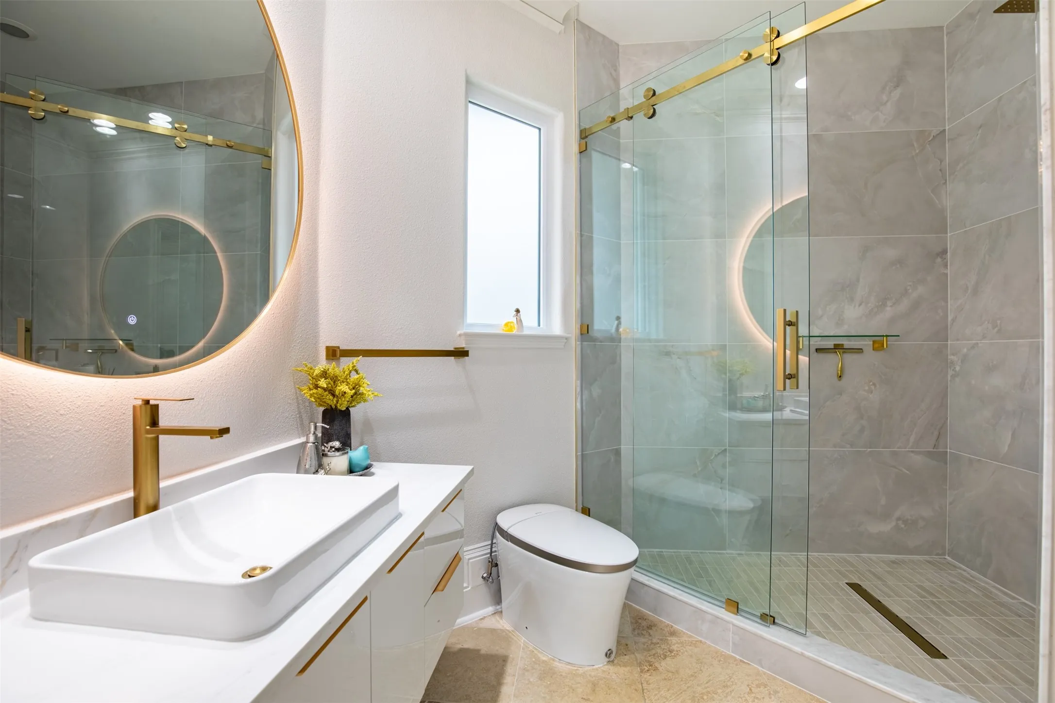 Bathroom featuring a frameless glass shower enclosure with gold-tone hardware, a rectangular vessel sink, a white floating vanity with gold-tone pulls, a round back-lit mirror, and grey tile shower walls