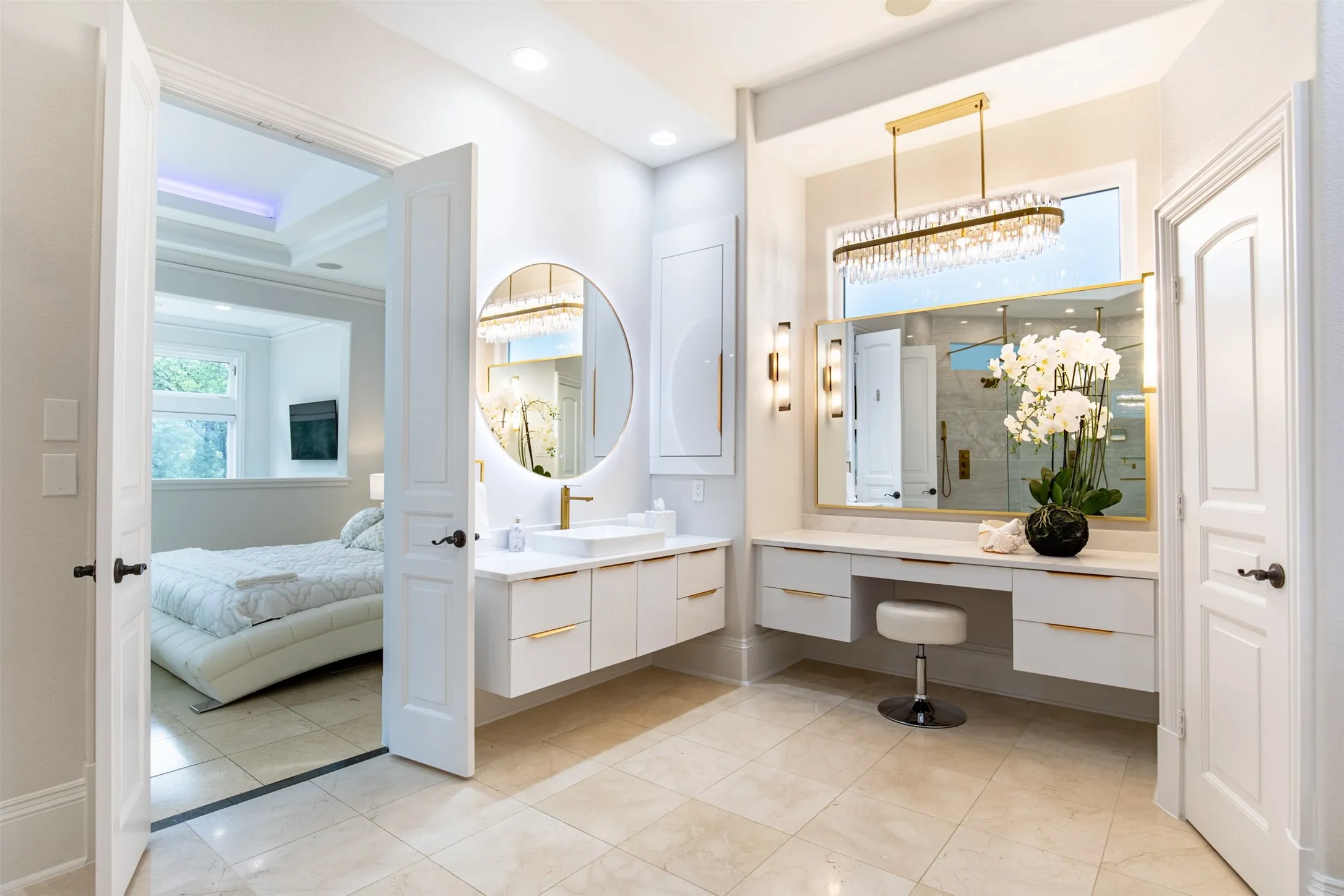 Spacious bathroom featuring light-toned tile flooring, a floating double vanity with a vessel sink, a large back-lit circular mirror, a vanity area with built-in drawers, and a modern linear chandelier