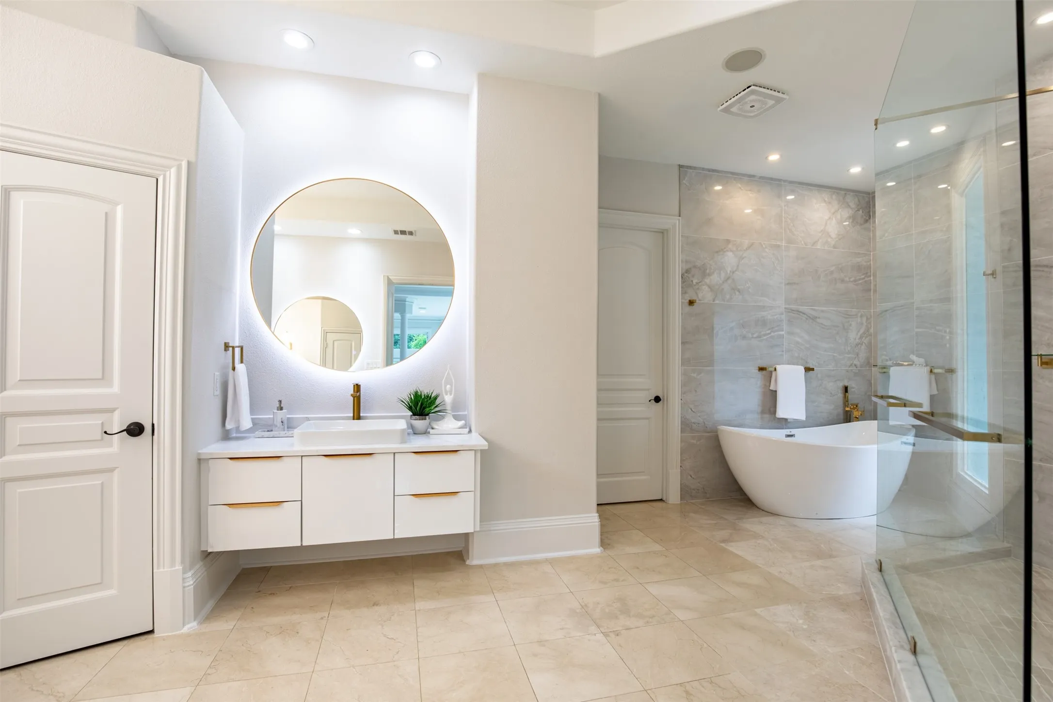 Spacious bathroom featuring light-colored tile flooring, a floating vanity with a vessel sink and illuminated round mirror, a separate freestanding soaking tub, and a glass-enclosed shower with large format wall tiling