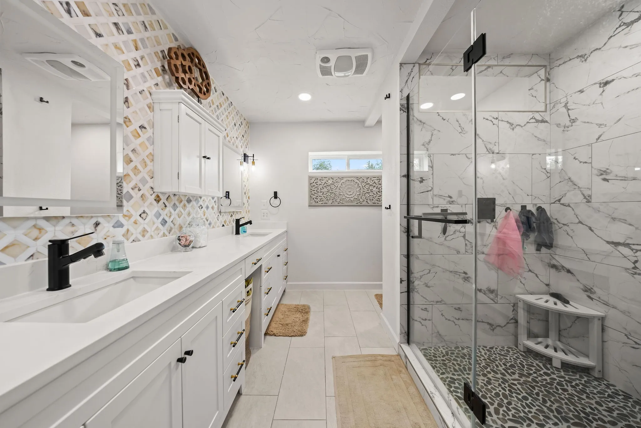 Spacious bathroom featuring a dual vanity with white cabinetry and countertops, a large glass-enclosed shower with pebble flooring, floor-to-ceiling marble-look tile, and a decorative accent wall with patterned tile