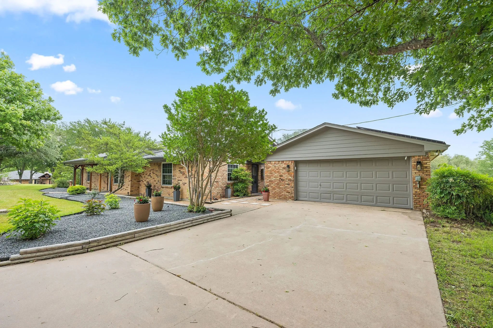 Brick exterior with a covered porch, an attached garage featuring a paneled door, a concrete driveway, and a landscaped front yard with a dark stone ground cover