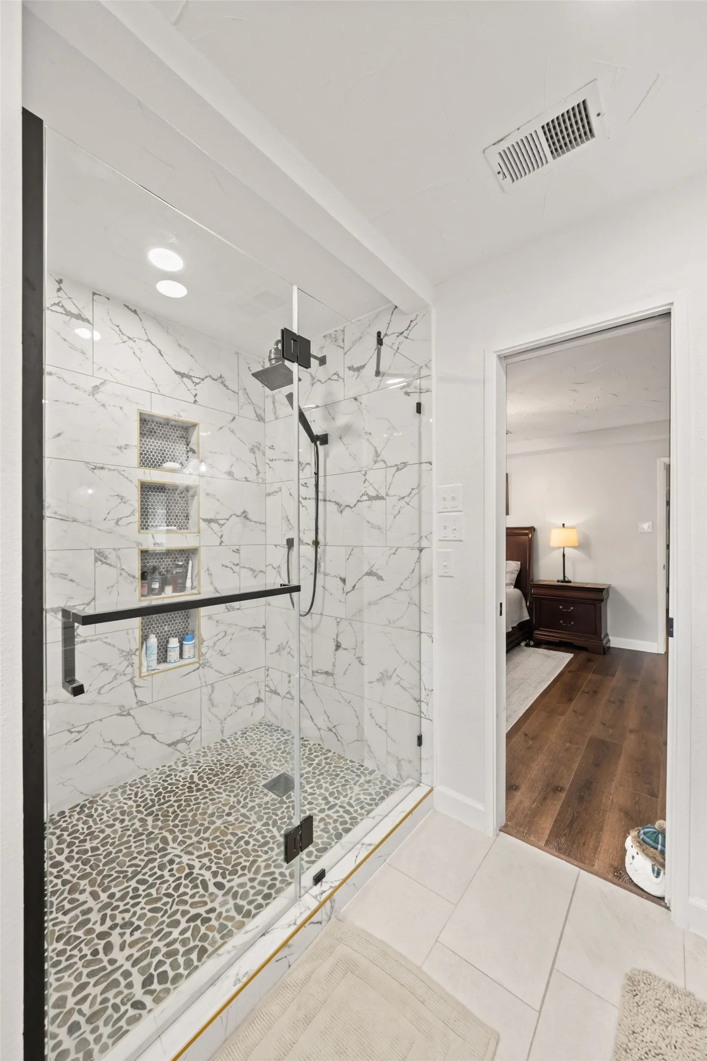 Walk-in shower with clear glass enclosure and black hardware, featuring marble-patterned tile walls, a pebble-stone floor, and recessed shower niches