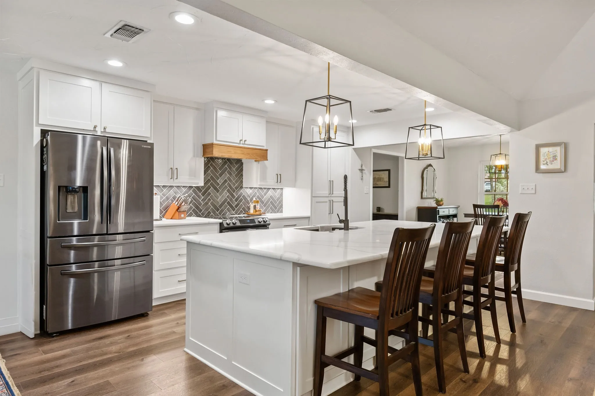 Modern kitchen featuring a large central island with seating, white shaker cabinetry, stainless steel appliances, herringbone tile backsplash, and wood-finish flooring