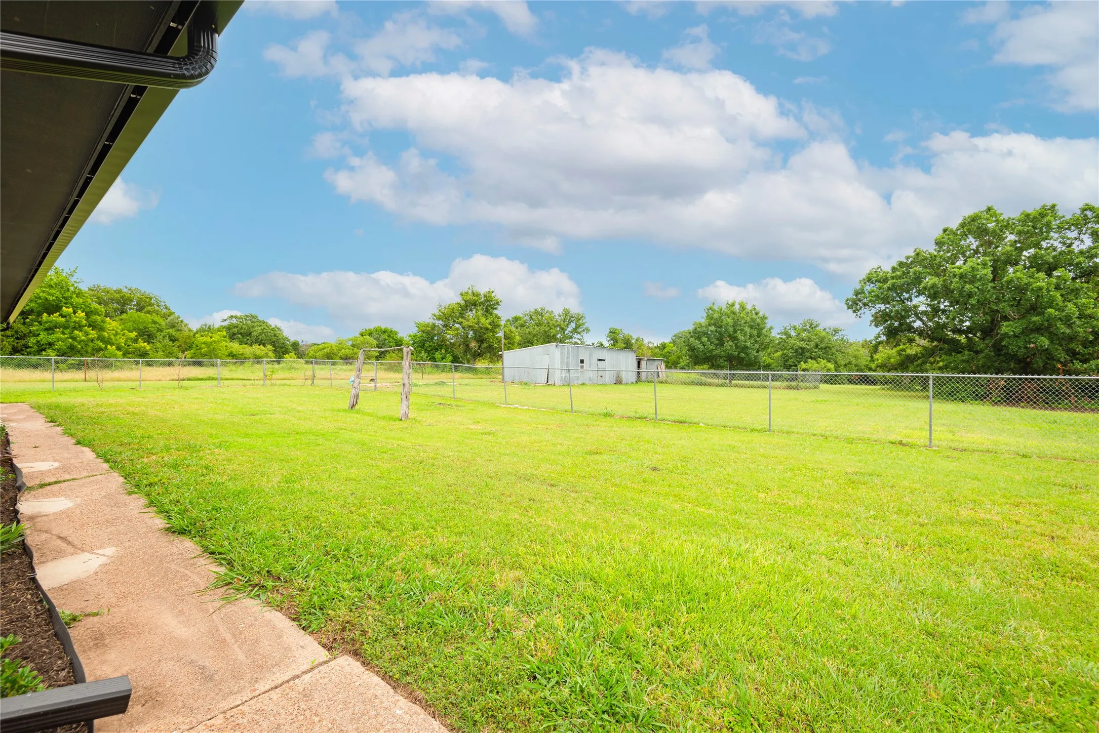 Expansive grassy yard with chain-link fencing, mature trees, a detached storage building, and a concrete pathway along the structure