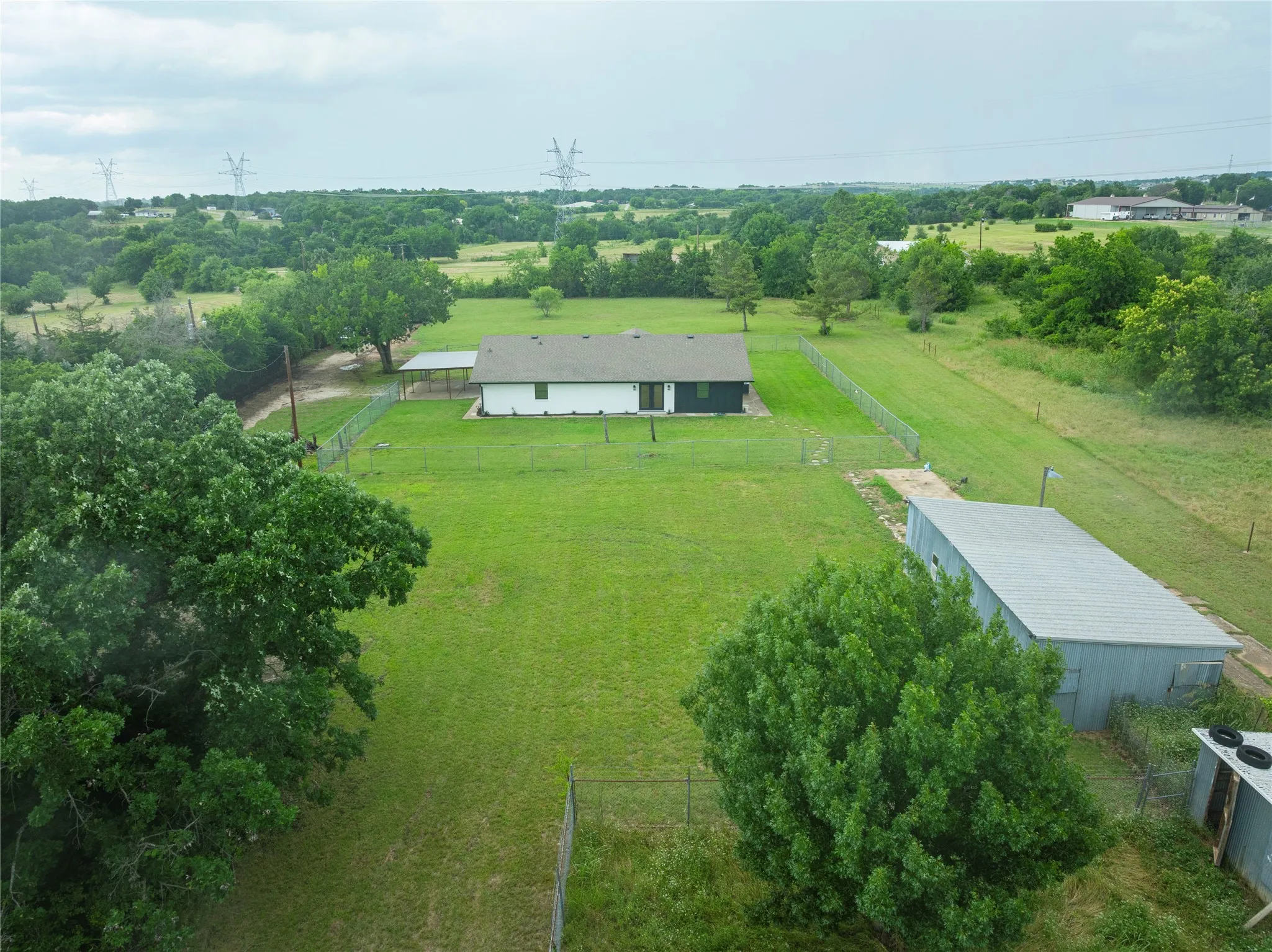 Expansive green property featuring a white residence with dark trim, a detached metal-sided outbuilding with a corrugated roof, and mature trees