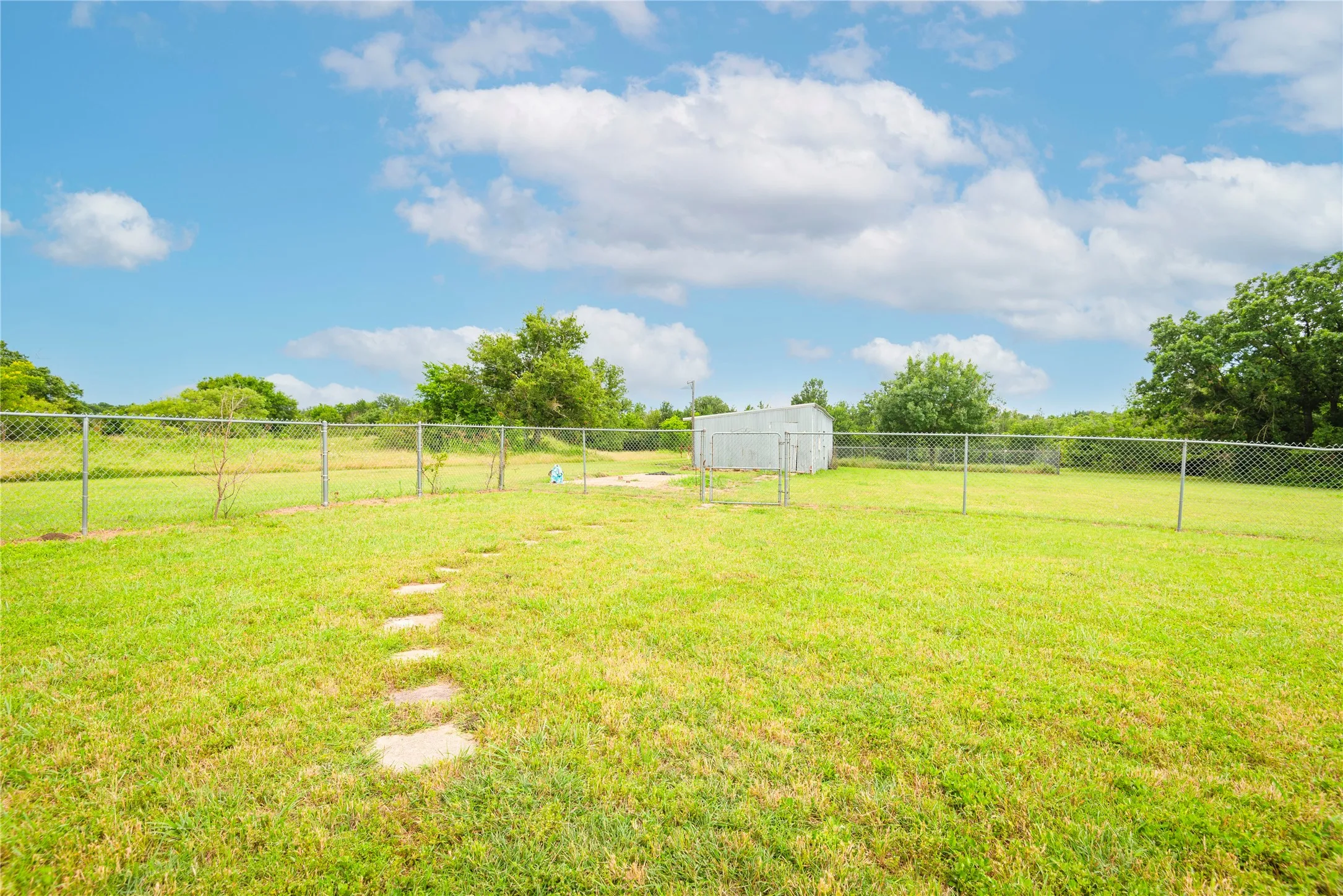 Expansive green lawn with a stepping stone pathway, chain-link fencing, mature trees, and a utility shed