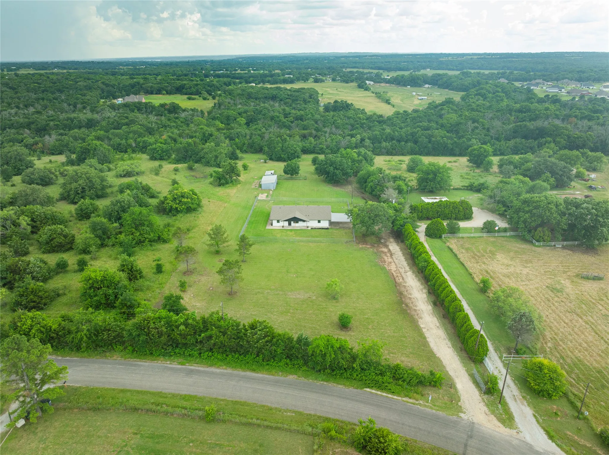 Expansive property featuring a single-story residence with a dark roof, a long gravel driveway, a secondary paved entrance, dense surrounding tree lines, and open green spaces