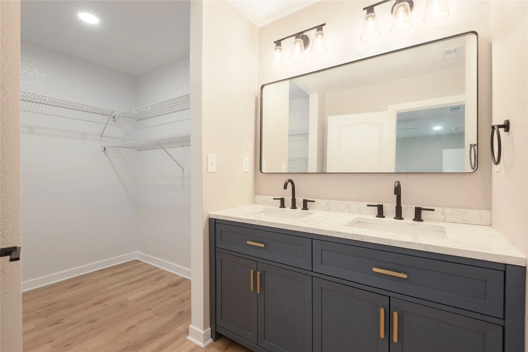 Dual vanity with undermount sinks and matte black fixtures, stone-look countertop, dark blue cabinetry with brass-toned hardware, large rectangular mirror, and an adjacent walk-in closet with wire shelving