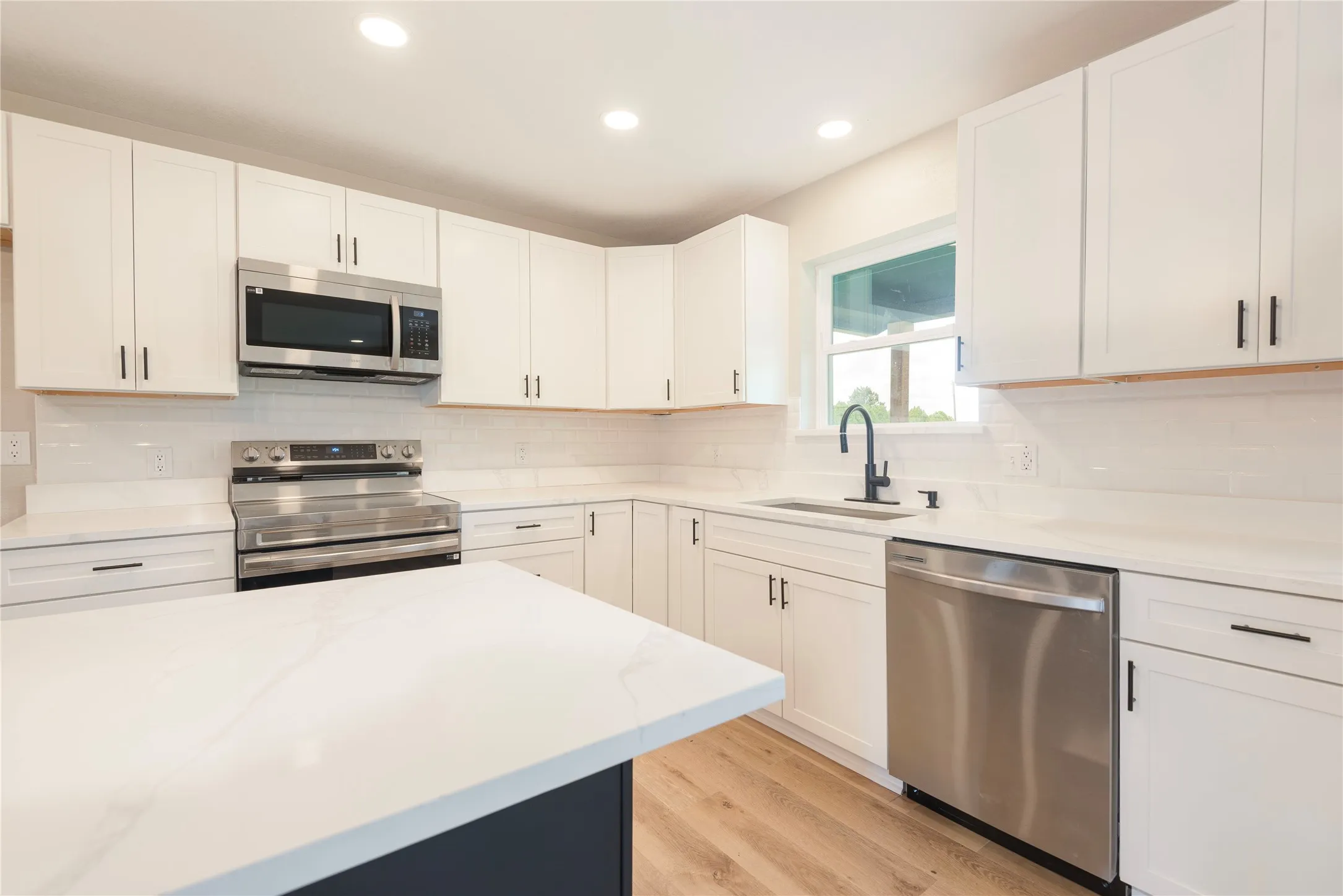 Modern kitchen featuring a central island with a light-toned countertop, white cabinetry with black hardware, stainless steel appliances, and wood-finish flooring