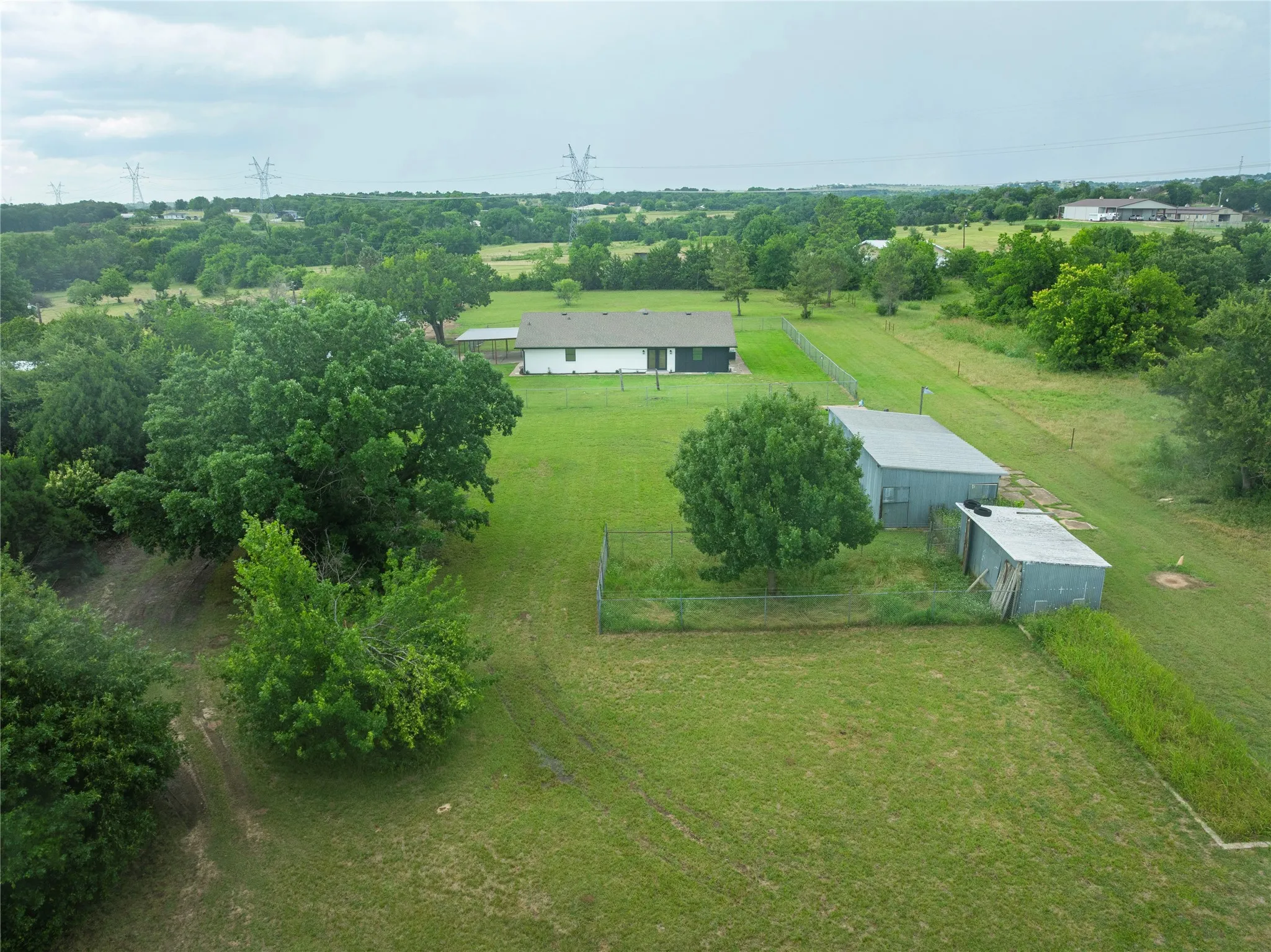 Expansive green landscape featuring a single-story residence with a dark roof and white siding, complemented by mature trees, two metal-sided outbuildings, and perimeter fencing